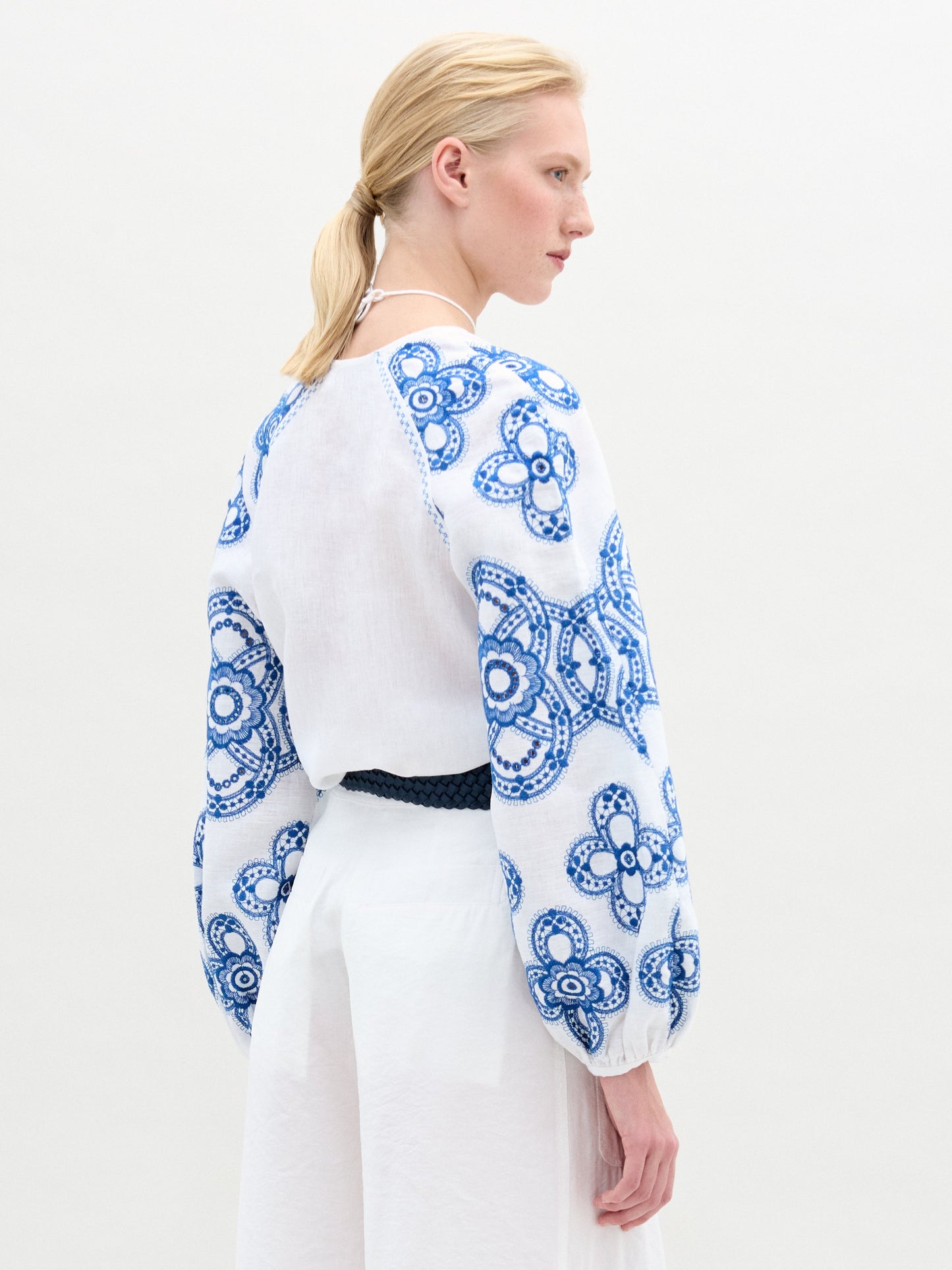 A woman with blonde hair in a low ponytail wears the Molveno Blouse—a linen top with blue floral embroidery and gathered sleeves—paired with a white skirt and dark belt, standing against a plain light background.