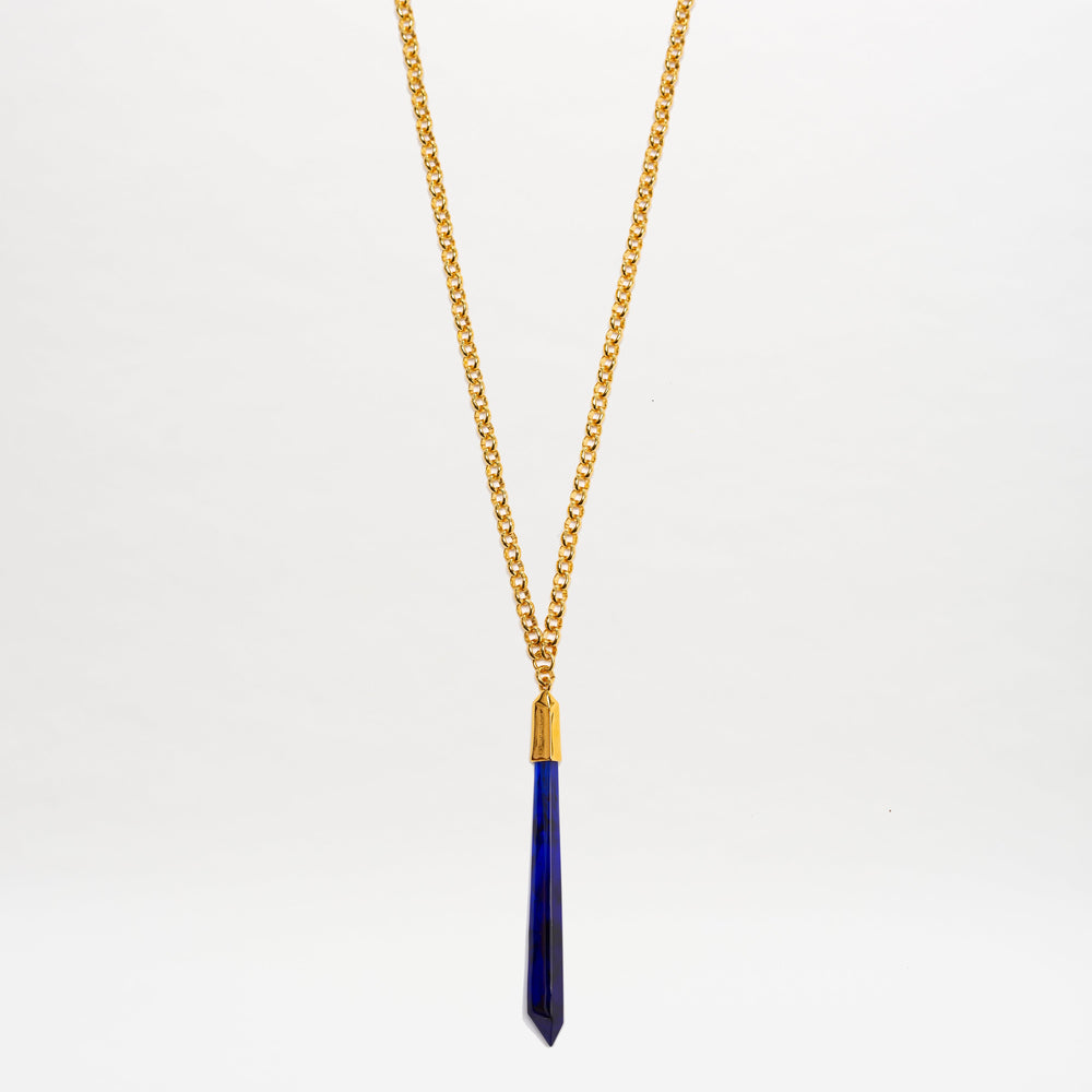 The Montse Necklace by SVT DESIGNS features a gold chain with a long, narrow dark blue geometric crystal pendant, shown hanging vertically against a plain white background.