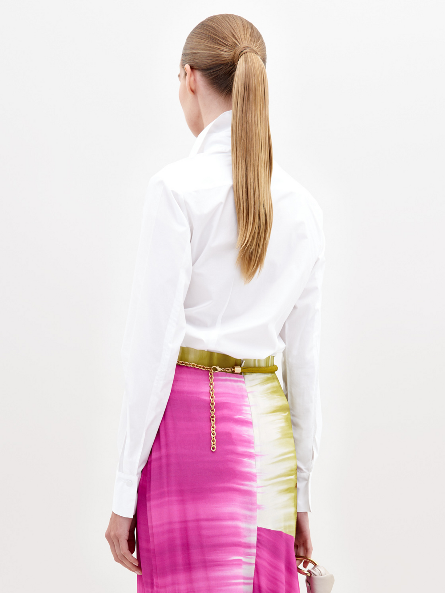 A woman with long, straight hair in a low ponytail faces away, wearing the Mercury Blouse—a 100% cotton white shirt—and a pink, white, and green watercolor-patterned skirt with a gold chain belt, holding a small beige bag.