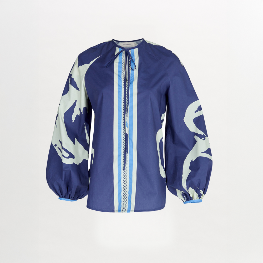 The Molveno Blouse by Silvia Tcherassi is a loose-fit, long-sleeved top in blue with white abstract sleeve patterns, two front vertical stripes, and a round neckline with tie detail—perfect for effortless everyday wear.