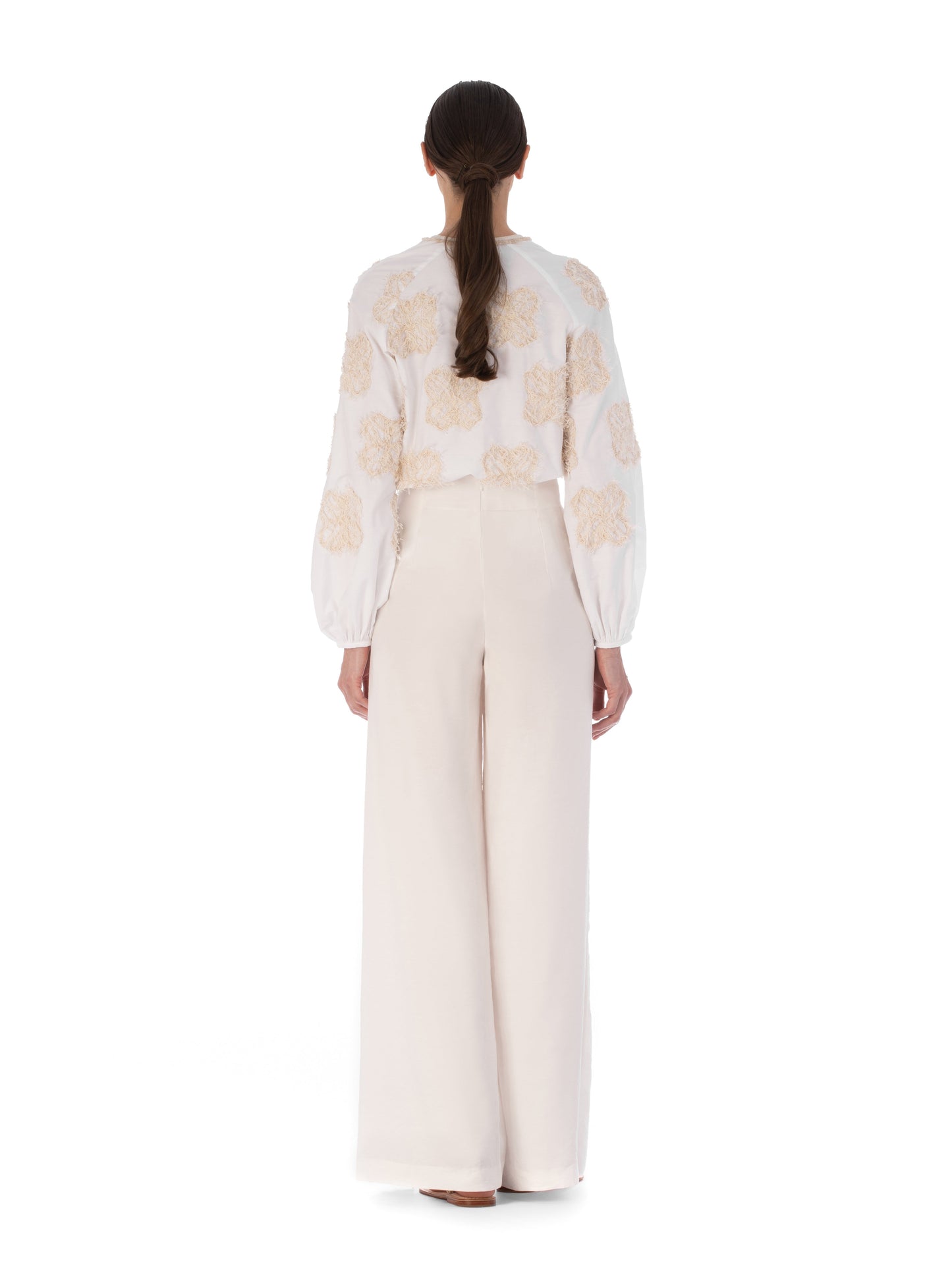 A person with long brown hair stands facing away, wearing the SVT DESIGNS Molveno Blouse, featuring floral motifs and a V neckline, paired with high-waisted cream wide-leg trousers against a plain white background.
