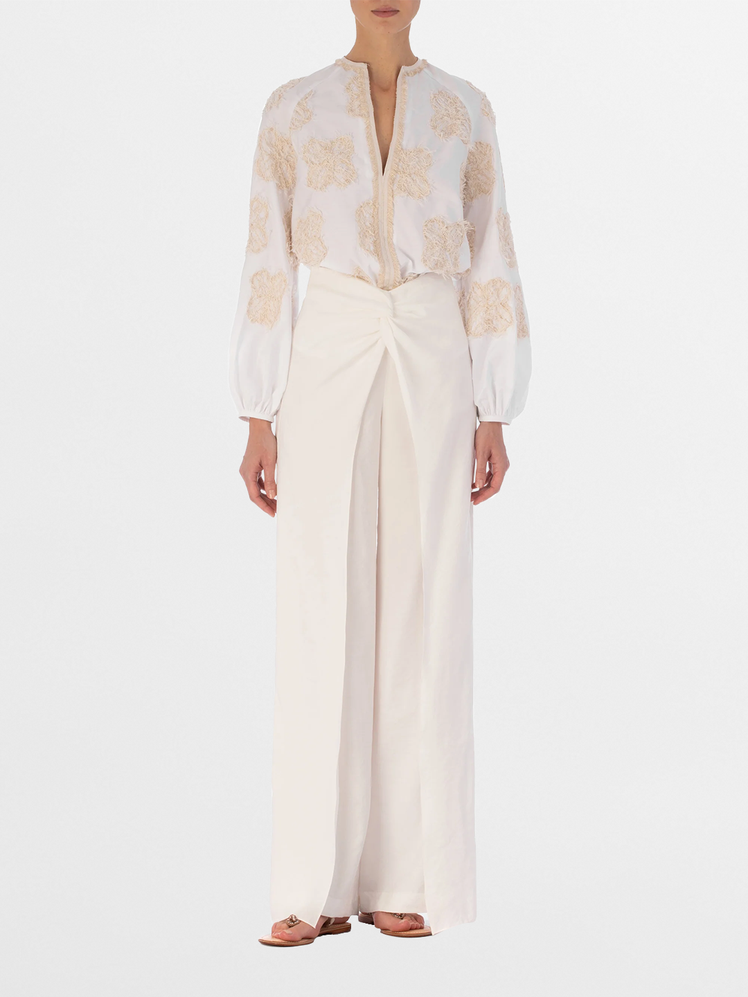 A person wearing the SVT DESIGNS Molveno Blouse—white cotton with beige floral embroidery and long sleeves—paired with high-waisted, wide-leg white pants and flat sandals, against a plain white background.