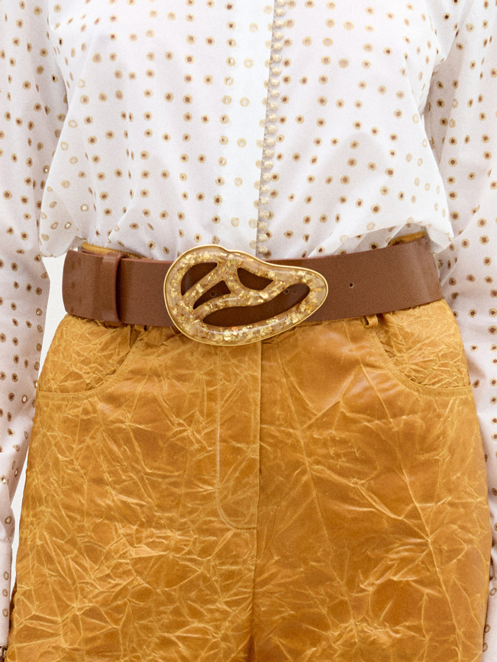 A person wearing a white shirt with gold dots, brown textured high-waisted pants, and the Nala Belt by SVT DESIGNS—an Italian leather belt with a large abstract Petralux buckle. Made in Colombia.