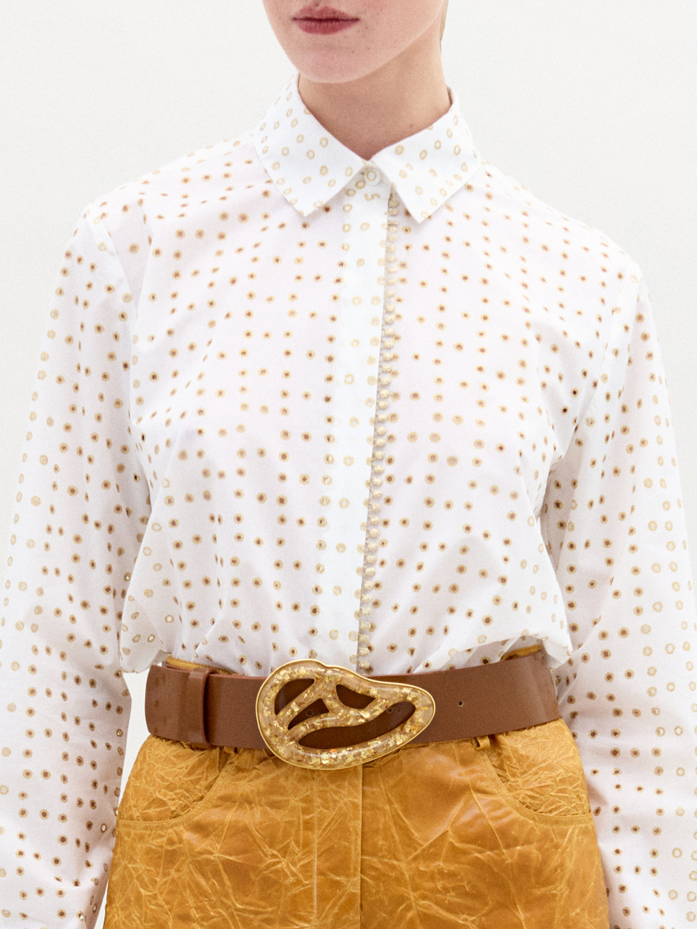 A person wears the SVT DESIGNS Naroa Blouse—a white long-sleeve top with a gold laser-cut dot design—tucked into high-waisted, textured mustard pants and finished with a wide brown belt featuring an abstract gold buckle.