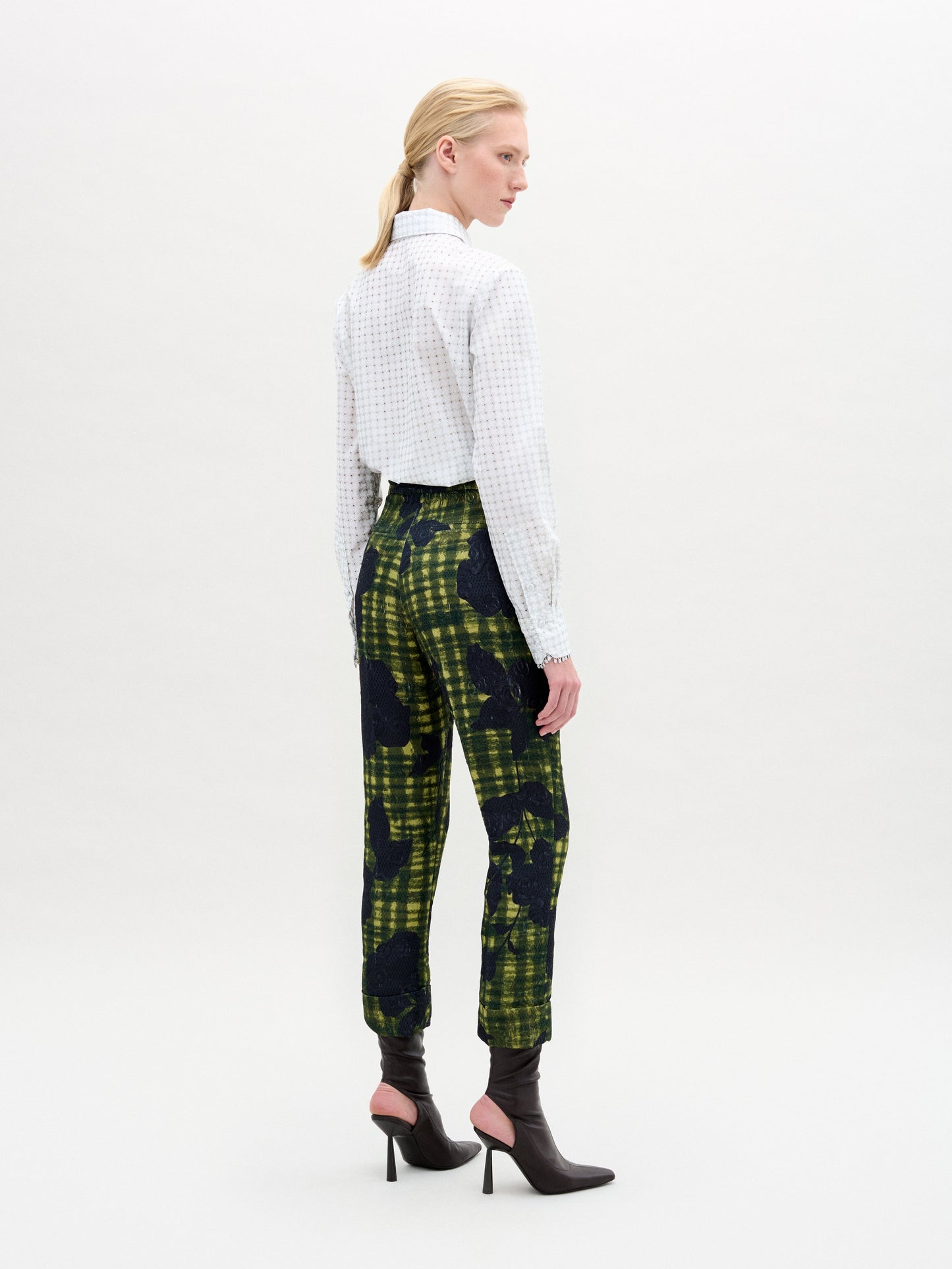 Against a neutral backdrop, a person models a chic outfit with the Naroa Blouse in Italian white patterned fabric, styled with green and blue patterned pants. The ensemble is completed by sleek black high-heeled boots.
