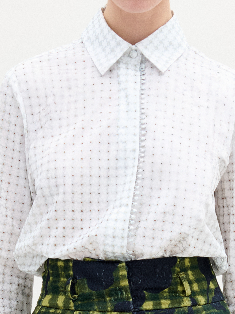 The individual is styled in SVT DESIGNS' Naroa Blouse, a long-sleeve white Italian fabric shirt with a subtle pattern and refined collar. Paired with high-waisted, dark-patterned green trousers, this ensemble exudes elegance while the individual's face remains unseen.