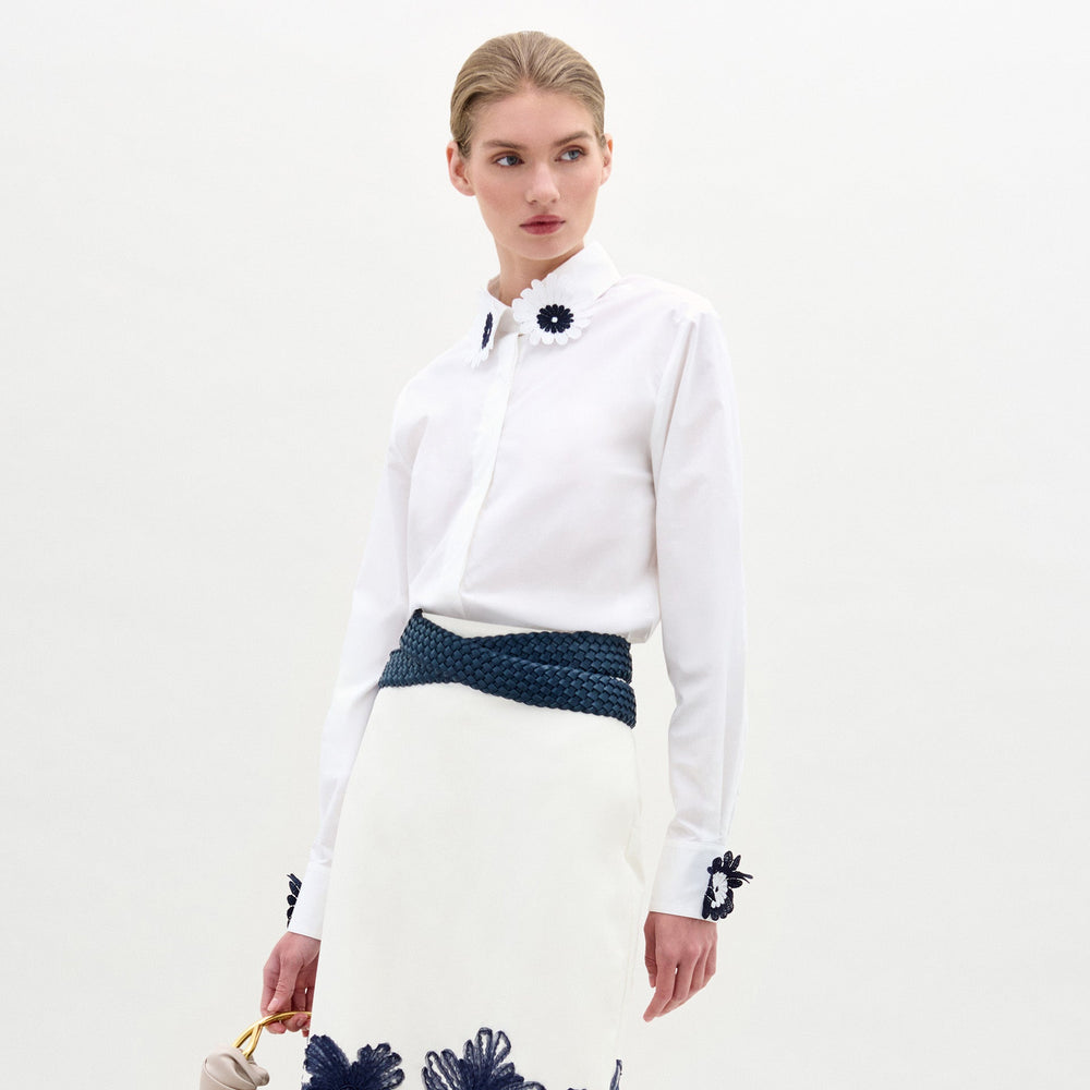 A woman stands against a plain white background wearing the Naroa Blouse by SVT DESIGNS, a long-sleeve Italian-fabric top with floral appliqué, paired with a white skirt with blue floral accents, a braided blue belt, and holding a light handbag.