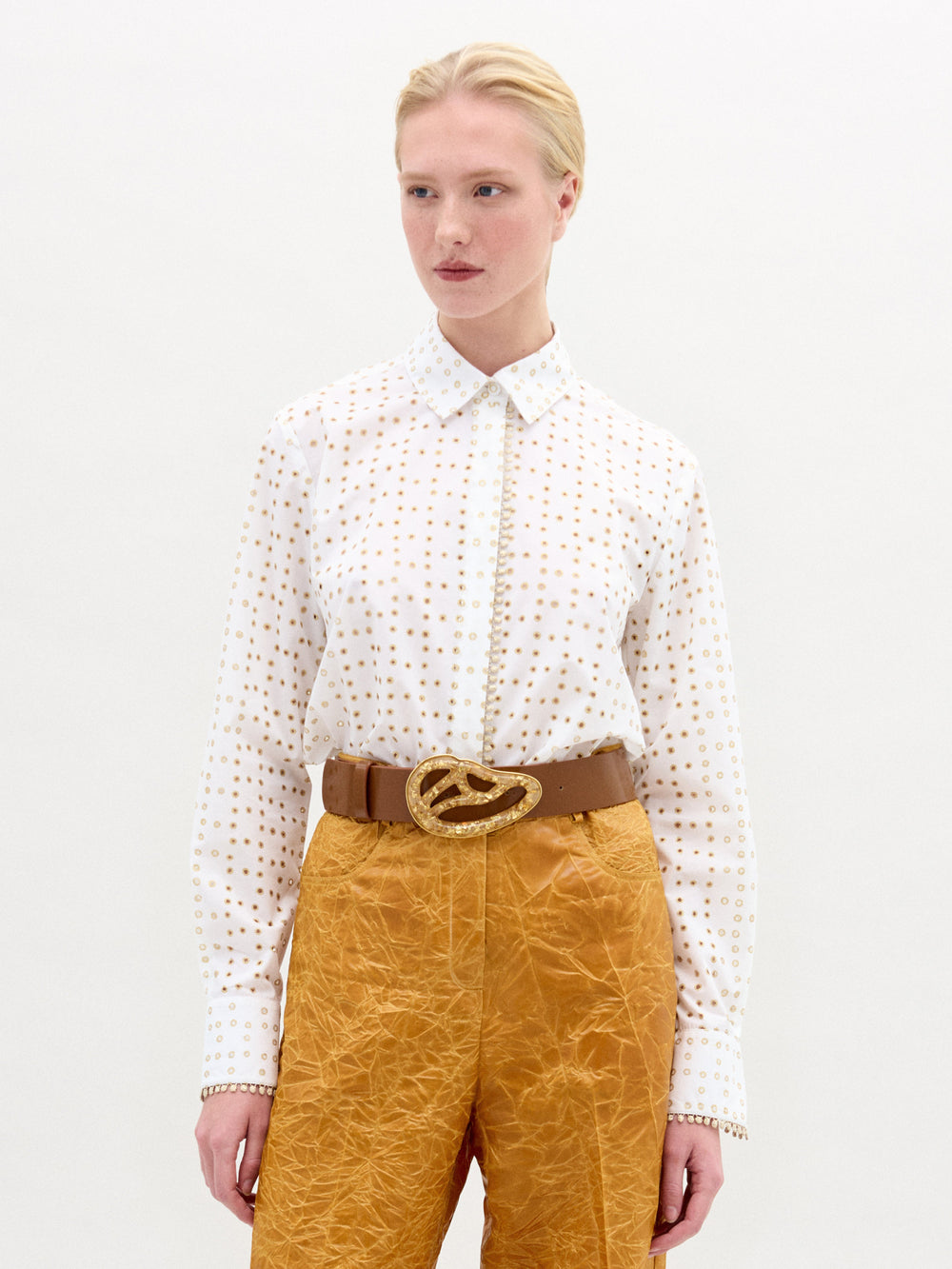 Against a plain light background, a person wears the SVT DESIGNS Naroa Blouse—white, long-sleeved with a laser-cut dot pattern—paired with mustard textured pants and a brown belt featuring a large gold buckle.
