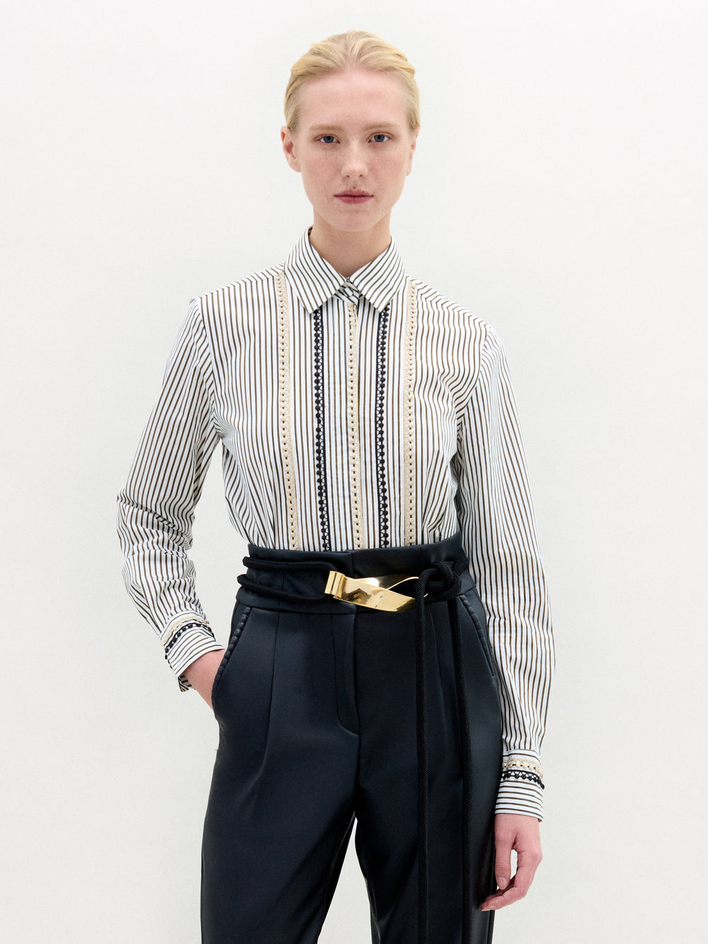A person stands against a plain white background, wearing the Naroa Blouse by SVT DESIGNS—a 100% cotton, vertically striped button-down shirt—paired with high-waisted dark trousers and a gold-buckled belt, one hand in their pocket.