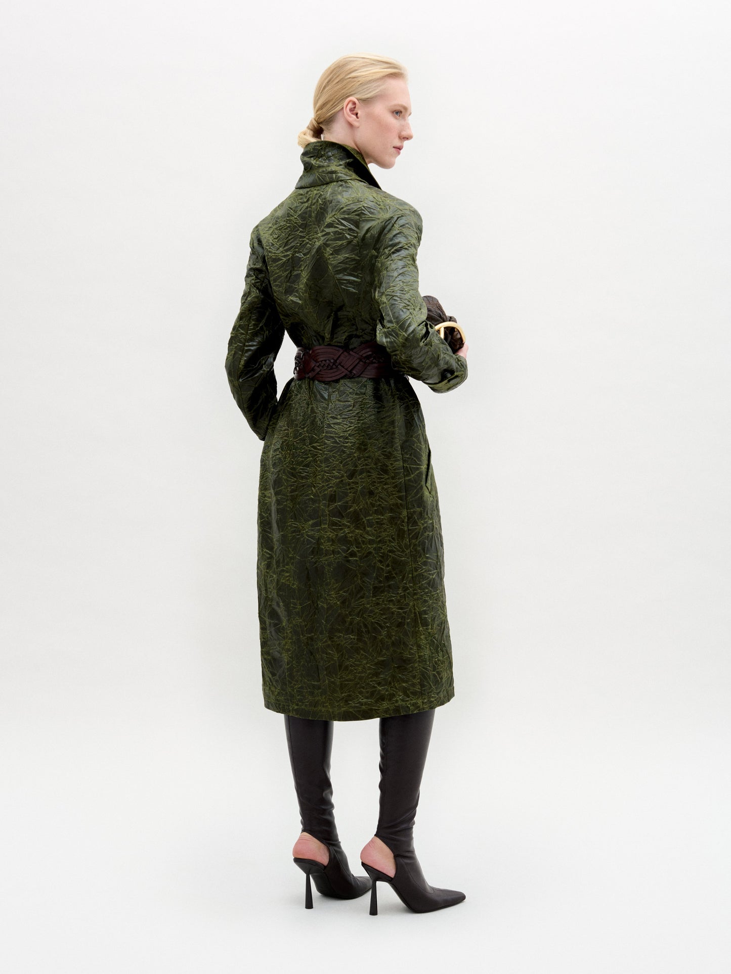 A woman wears the Nilo Trench, a textured dark green coat crafted from Italian fabric, paired with dark boots, a large gold belt buckle, and holding a small dark clutch against a plain background.
