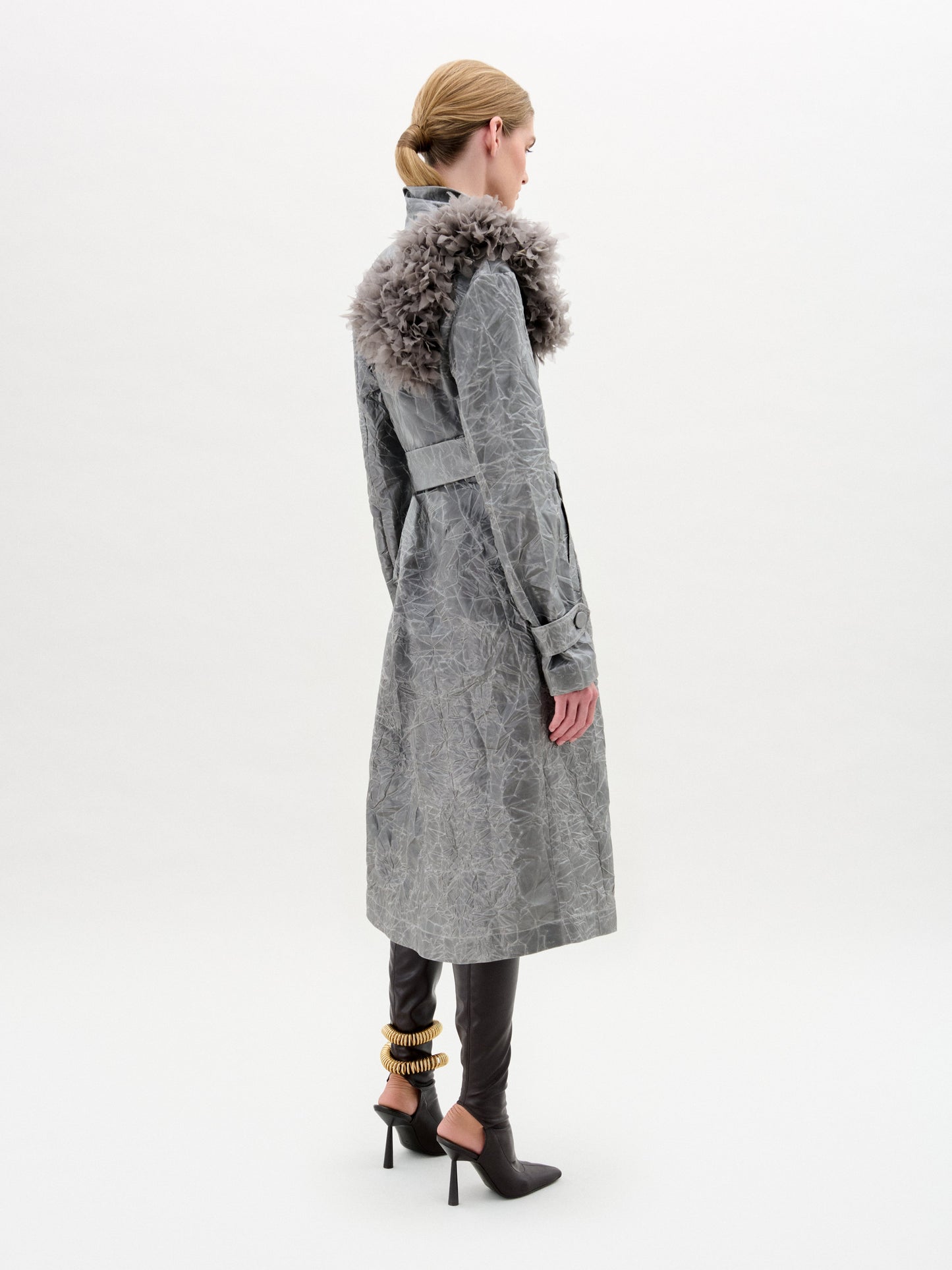 A woman stands against a plain background wearing the Nilo Trench, a textured gray structured coat with a large floral shoulder detail, paired with black heeled boots and a gold ankle accessory.