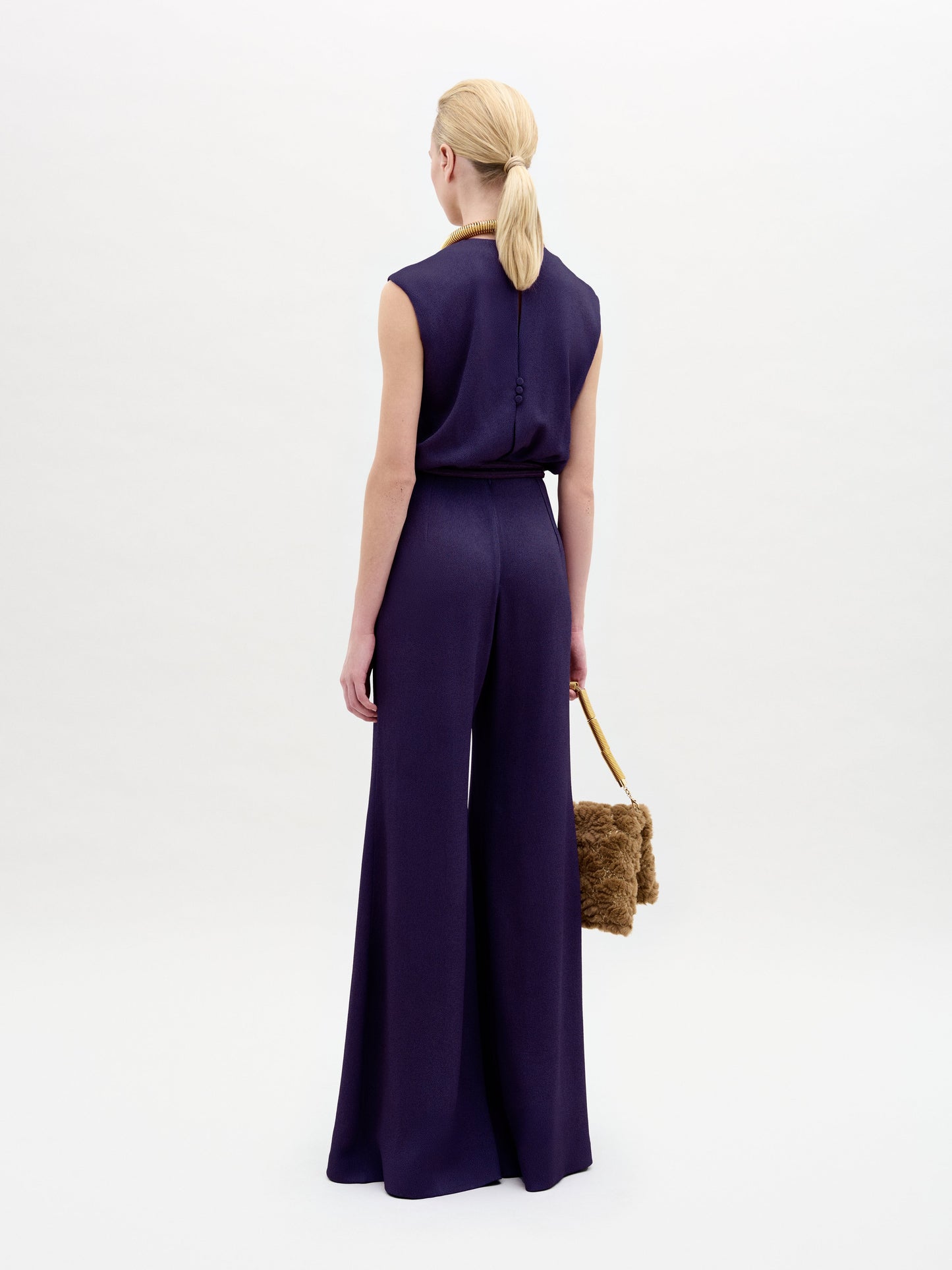 A woman with blonde hair in a low ponytail wears the sleeveless, wide-leg Noel Jumpsuit and holds a brown faux fur handbag, standing with her back to the camera against a plain white background.