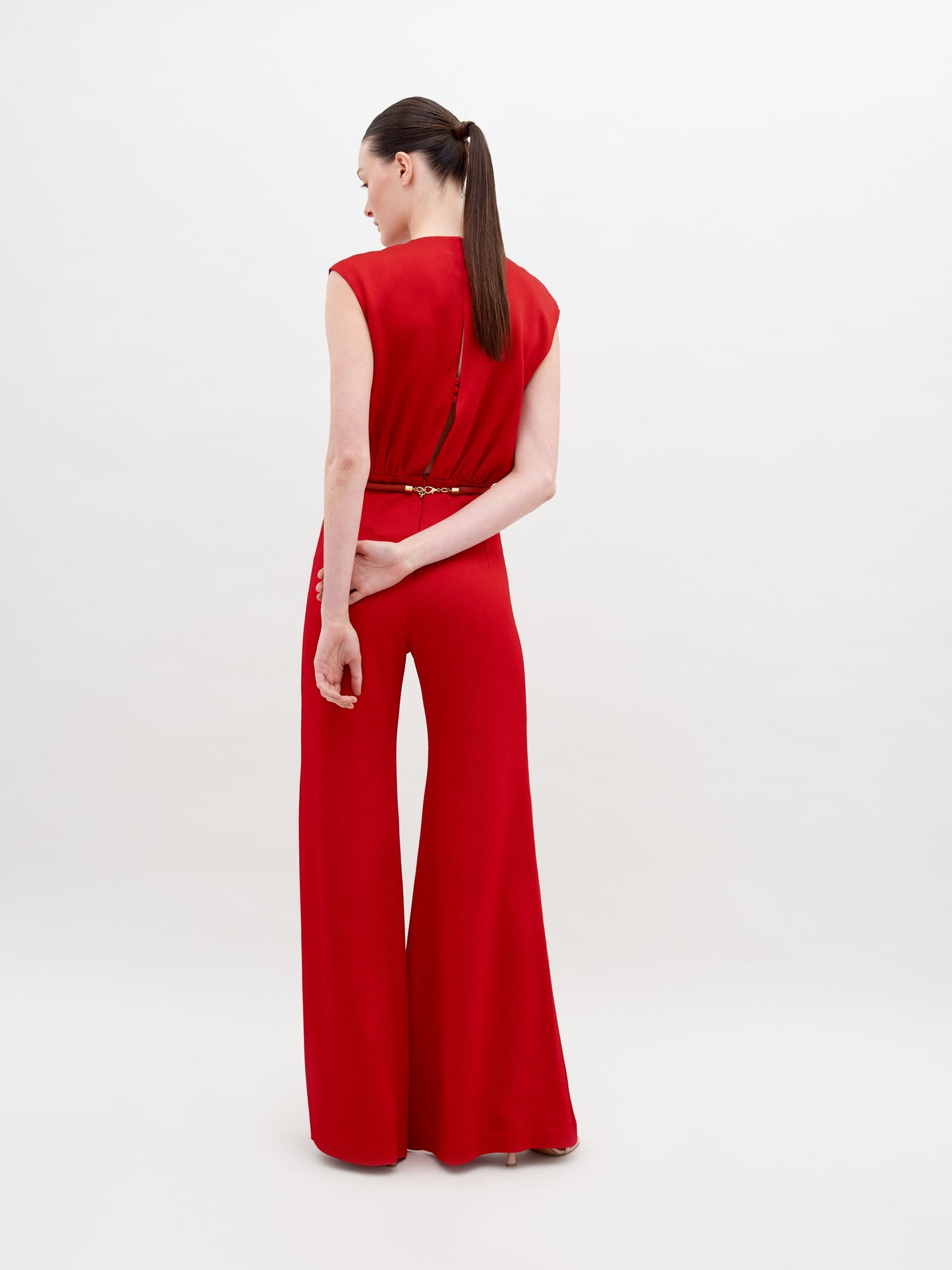 A woman stands facing away, wearing the Noel Jumpsuit in bright red, crafted from luxurious Italian textile, her hair in a low ponytail and one arm behind her back, set against a plain white background.