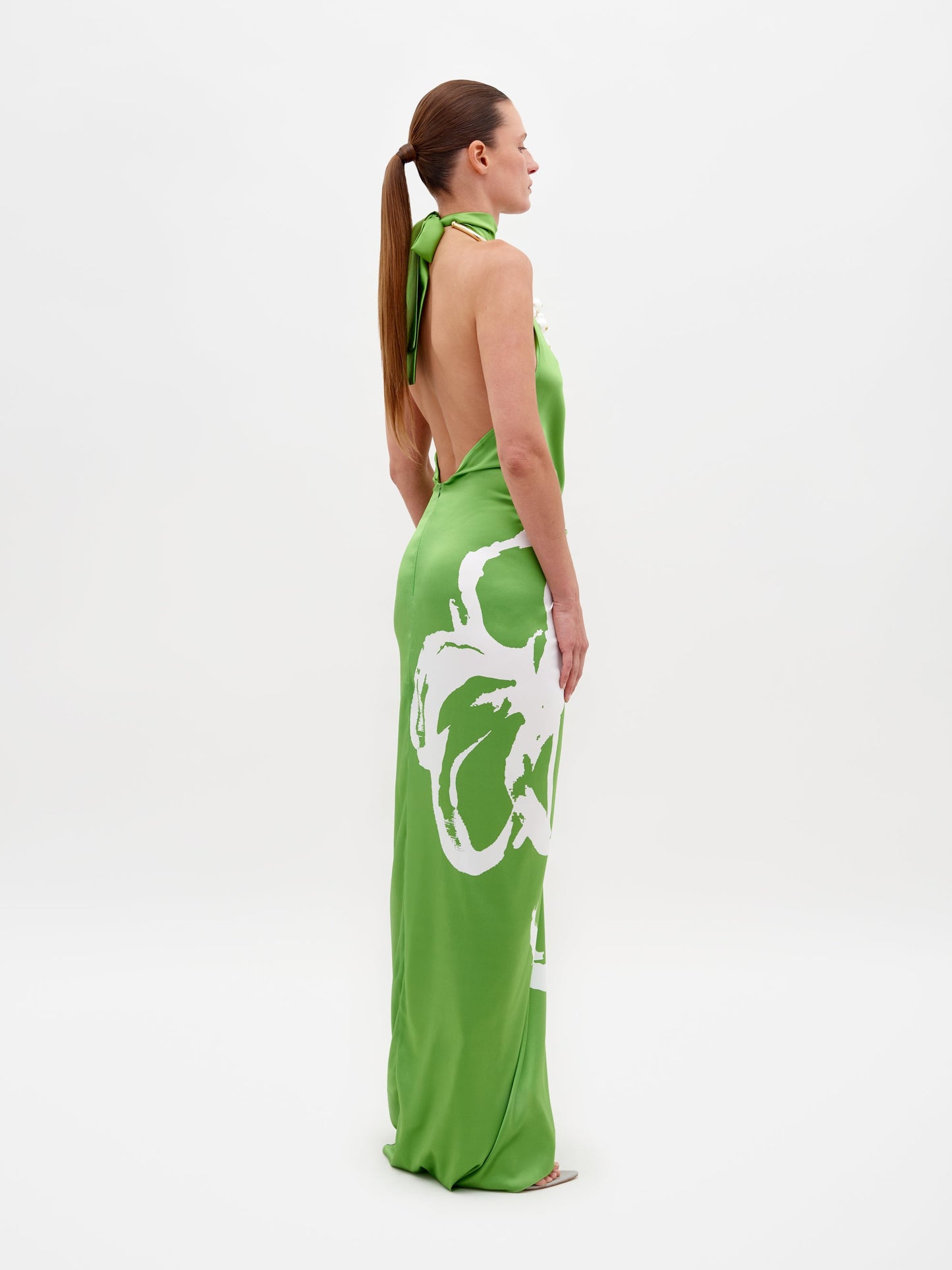 A woman poses in profile against a plain backdrop, wearing the Olatz Dress by SVT DESIGNS: a high-neck halter maxi silk dress with an abstract print of large white flowers on green. Her sleek ponytail complements the dress's elegance.