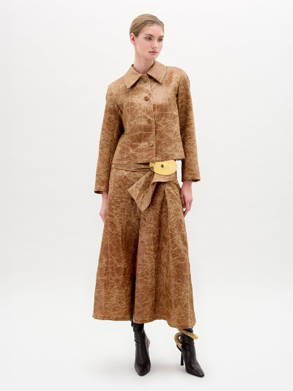 A person stands against a plain white background wearing the Olga Skirt by SVT DESIGNS, paired with a textured brown long-sleeve top, large buttons, black ankle boots, beige belt bag, and hair pulled back.