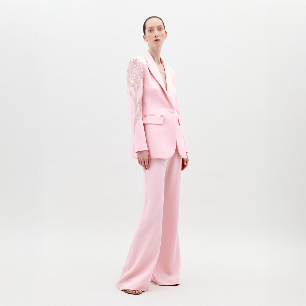 A person stands against a plain white background wearing the Palermo Pant by SVT DESIGNS, a tailored fit light pink pantsuit with high-waisted wide-leg trousers and a blazer featuring floral embroidery on the right shoulder.