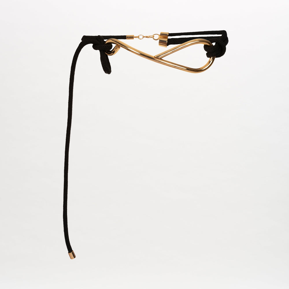 The Pat Belt by SVT DESIGNS features a gold-tone infinity buckle with black knotted cords—one strap hangs longer than the other—all set against a plain white background.