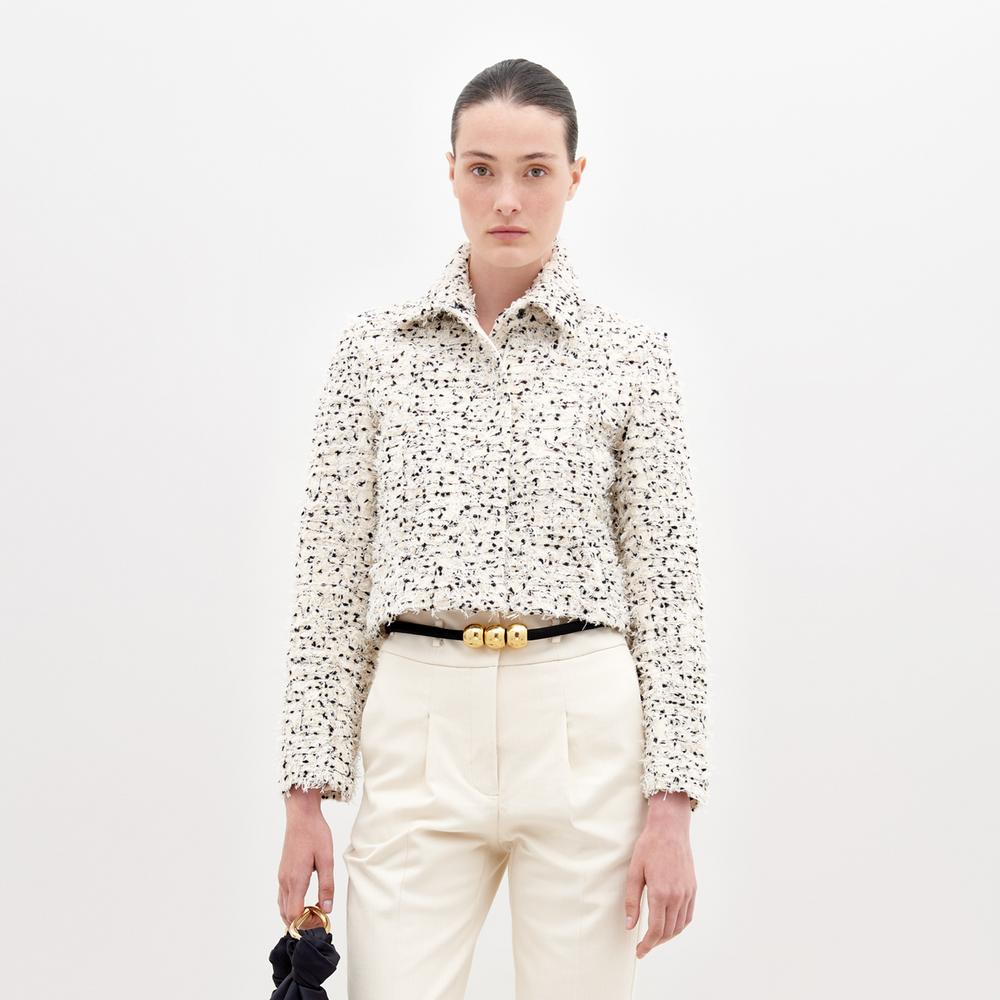 A woman poses against a white backdrop wearing the SVT DESIGNS Perla Jacket—a textured, long-sleeve cream blouse with black accents crafted from fine Italian textile—paired with cream high-waisted trousers, a black belt with gold accents, and holding a black item.