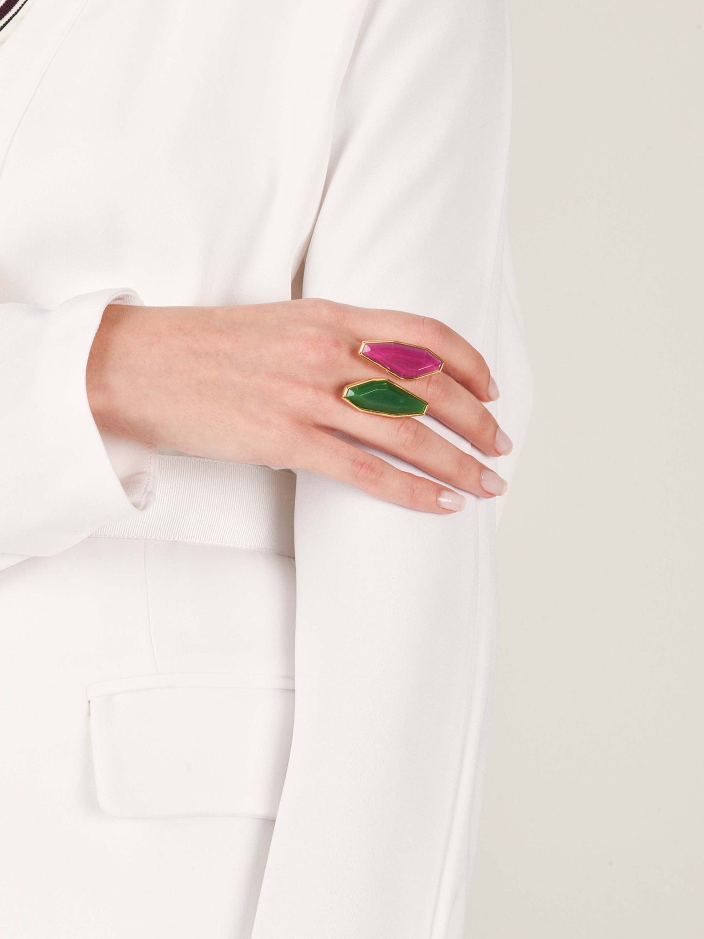 A person in a white blazer crosses their arm, showcasing the Suzy Ring Magenta Emerald—an asymmetrical gold band adorned with two large fantasy stones, one magenta and one emerald, against a plain light background.
