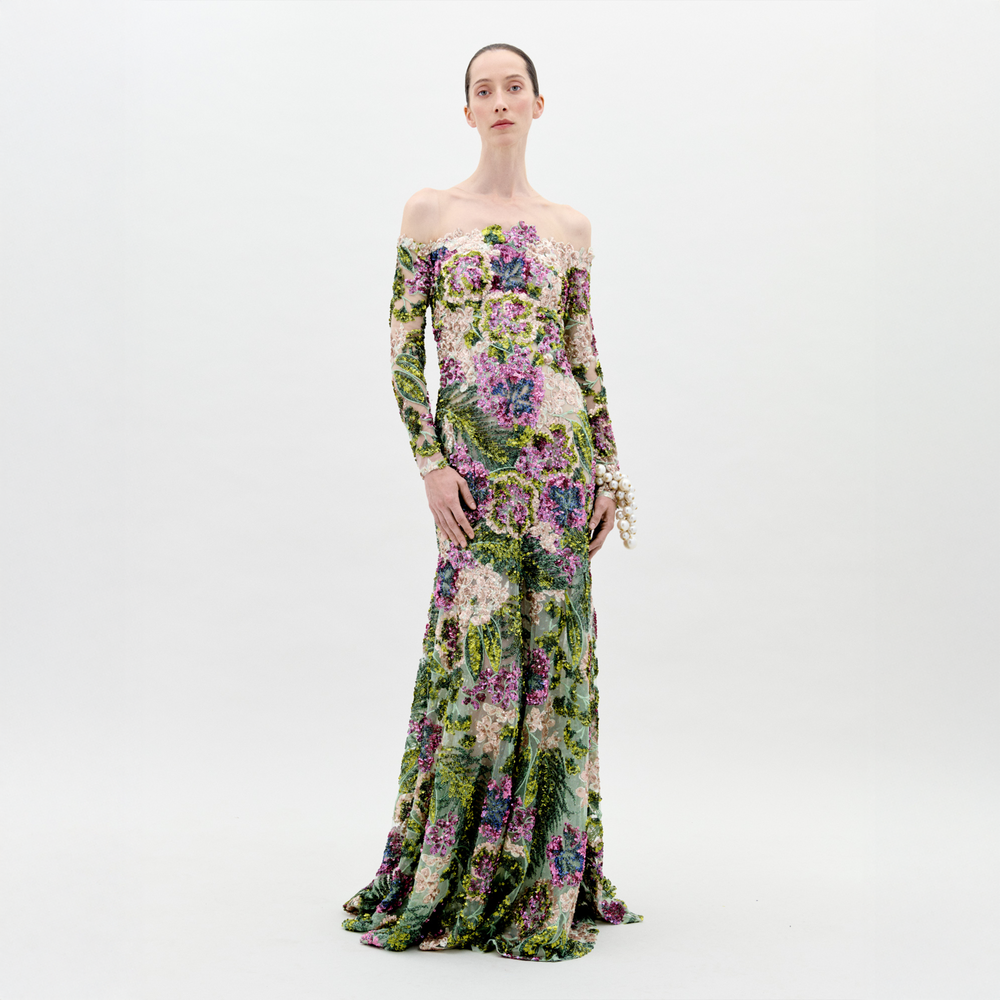 A woman stands against a white background wearing the Rafaella Dress by SVT DESIGNS—an off-the-shoulder, long-sleeved atelier piece with a fitted bodice and green, purple, and pink botanical print. Her hair is pulled back and she wears a pearl bracelet.