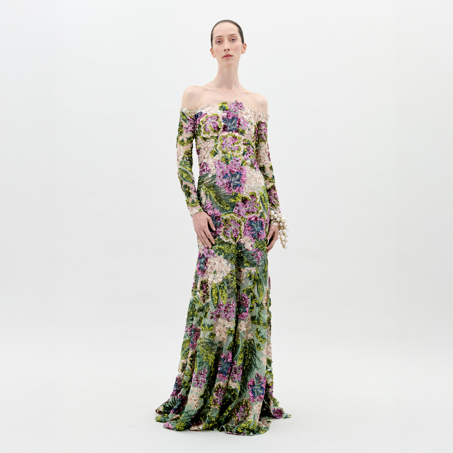 A woman stands against a white background wearing the Rafaella Dress by SVT DESIGNS—an off-the-shoulder, long-sleeved atelier piece with a fitted bodice and green, purple, and pink botanical print. Her hair is pulled back and she wears a pearl bracelet.