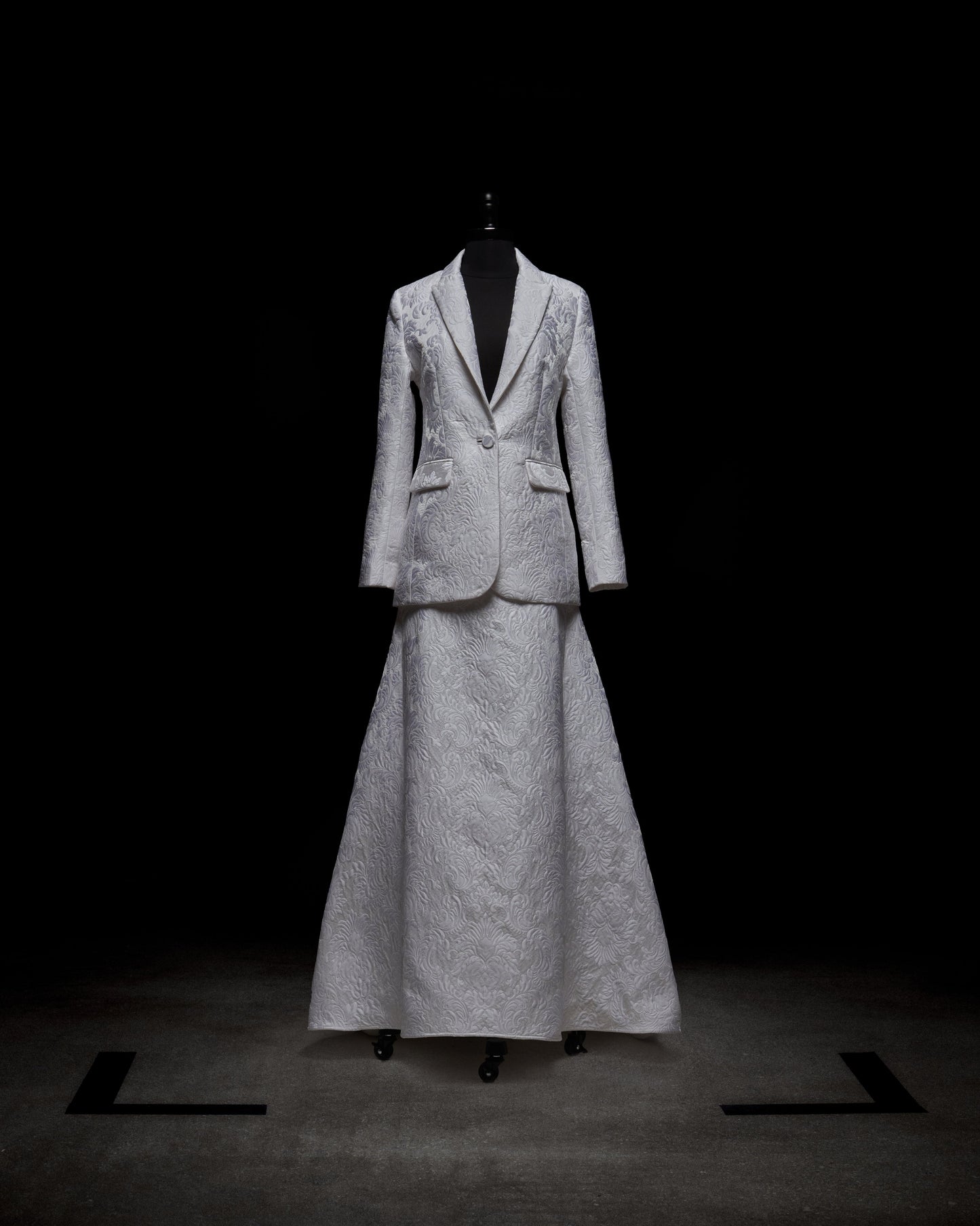 A mannequin features a chic ensemble from SVT DESIGNS, showcasing the 'Imola Skirt,' a long, textured white piece paired with an elegantly embroidered matching blazer against a dark backdrop.