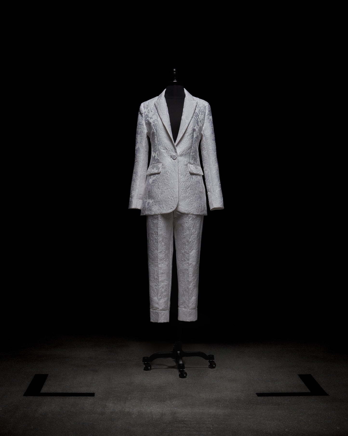 A silver patterned Rebeca Jacket by SVT DESIGNS is displayed on a mannequin against a black background, featuring a single-breasted style with notched lapels and matching trousers. The lighting accentuates the fabric's texture and intricate design.
