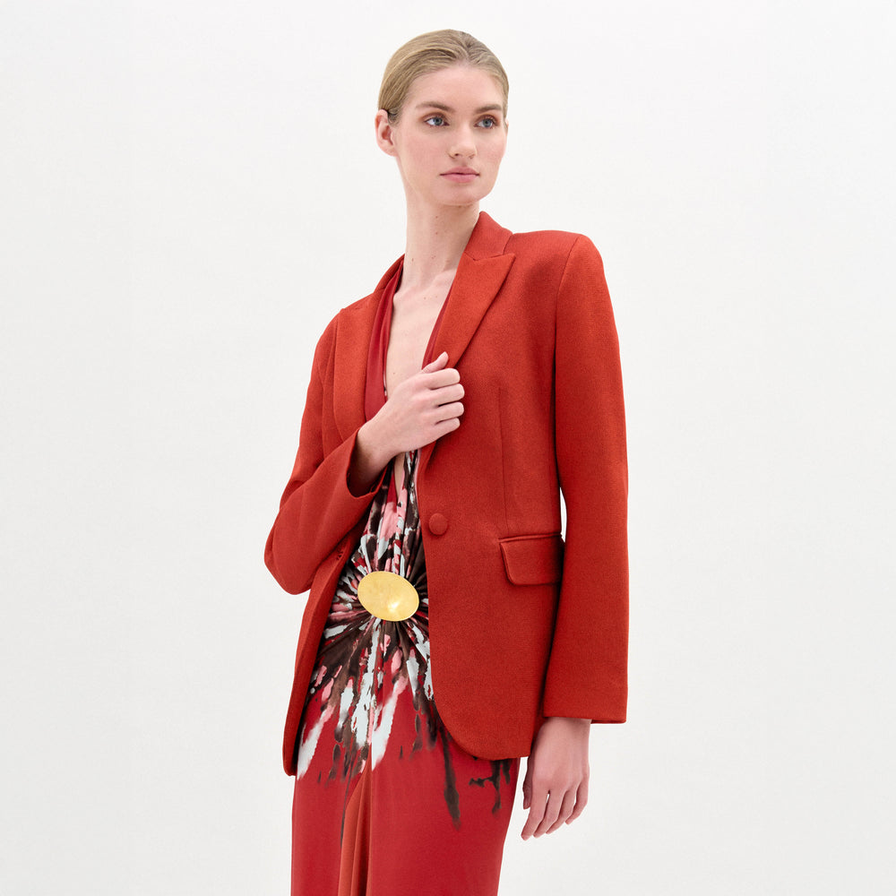 A person wears the fitted Rebeca Jacket by SVT DESIGNS over a patterned red, white, and brown dress with a large gold circular belt buckle. Their light hair is pulled back as they look to the side against a plain background.