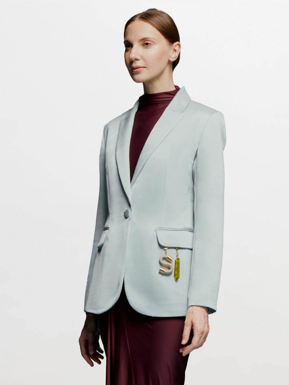A woman faces slightly left against a white background, wearing the SVT DESIGNS Rebeca Jacket—a relaxed fit light blue crepe blazer—over a burgundy dress. A gold "S" keychain hangs from the jacket's pocket.
