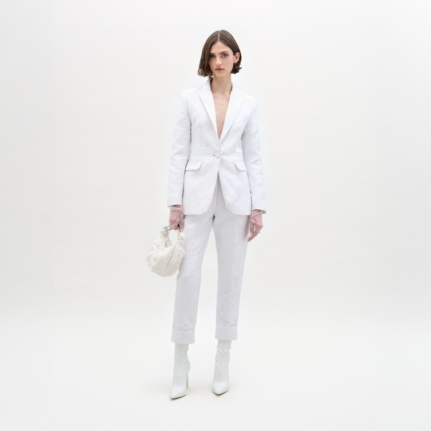 A person with short brown hair and a neutral expression stands against a white background, wearing the SVT DESIGNS Moad Pant—textured white, straight-leg Italian fabric—paired with white boots and a small white handbag.