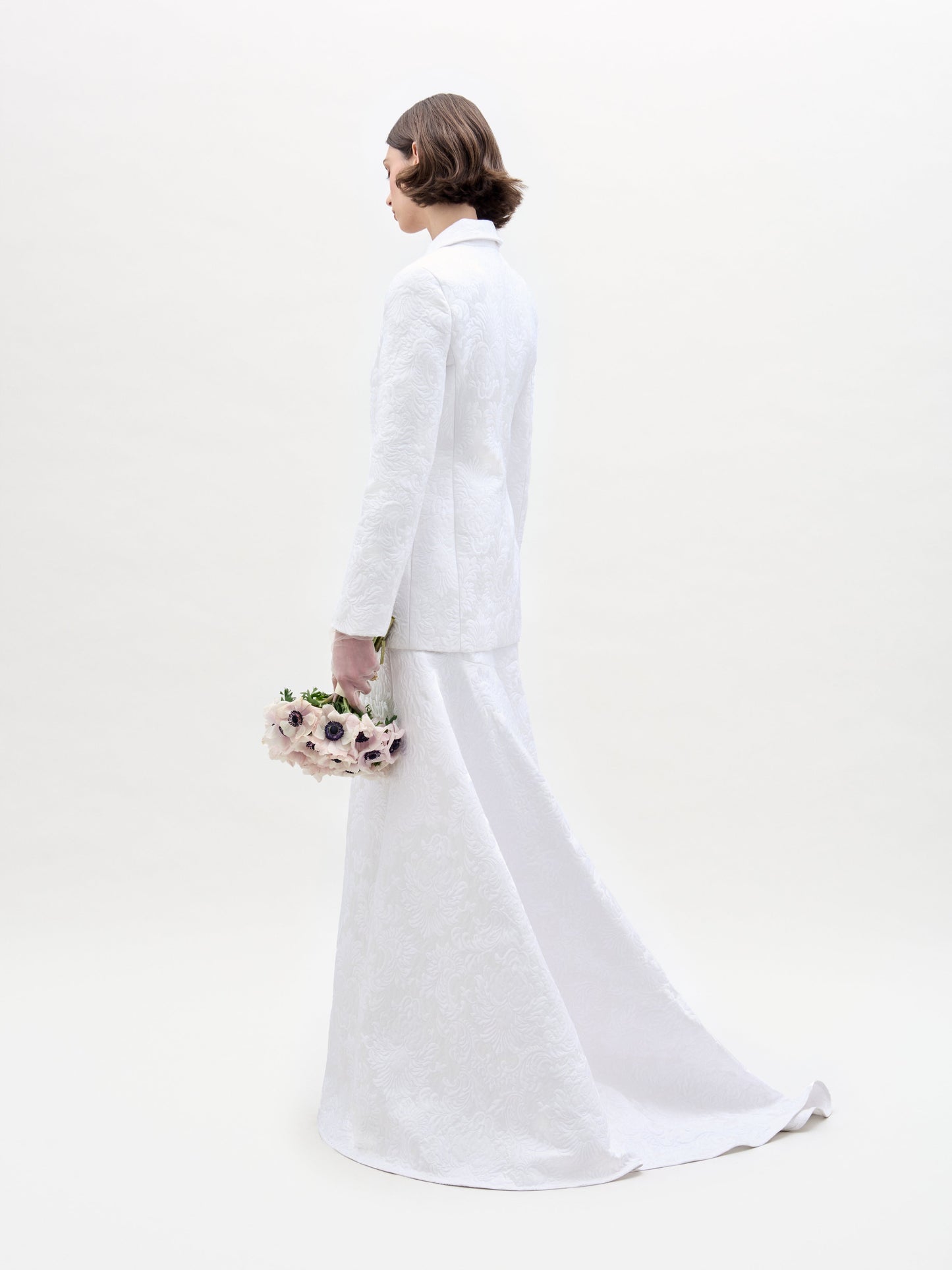 A person with short brown hair faces away from the camera, dressed in a white textured suit featuring the Imola Skirt by SVT DESIGNS. They hold a bouquet of light-colored flowers with dark centers against a plain white backdrop.