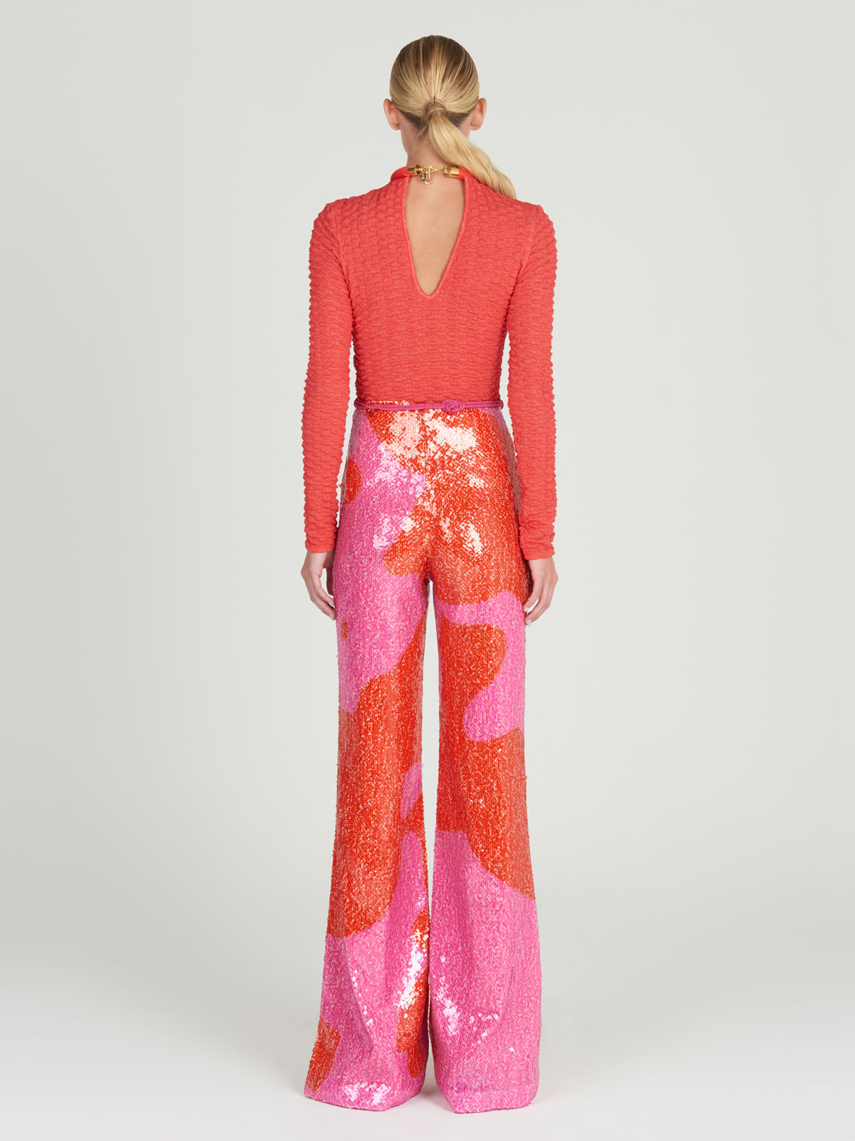 A woman with blonde hair in a low ponytail faces away, wearing the Jari Top—an embossed jacquard red long-sleeve with a keyhole back—paired with high-waisted sequined wide-leg pants in bold pink and orange abstract patterns.