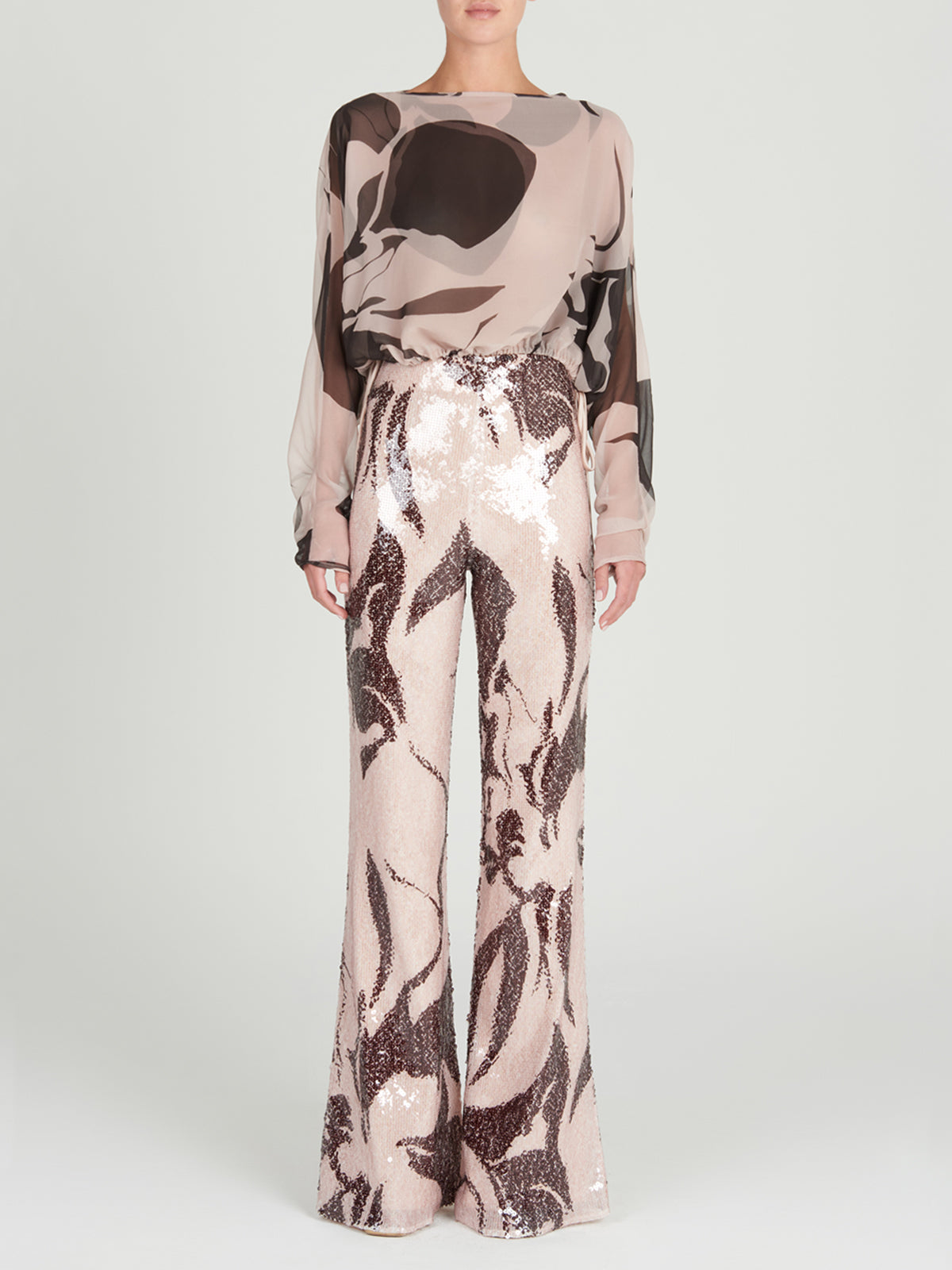 A person stands facing forward in the Silvia Tcherassi Macerata Blouse, a lightweight silk chiffon, sheer long-sleeve top with an abstract pattern, paired with high-waisted, wide-leg sequin pants featuring a dark, swirling design.
