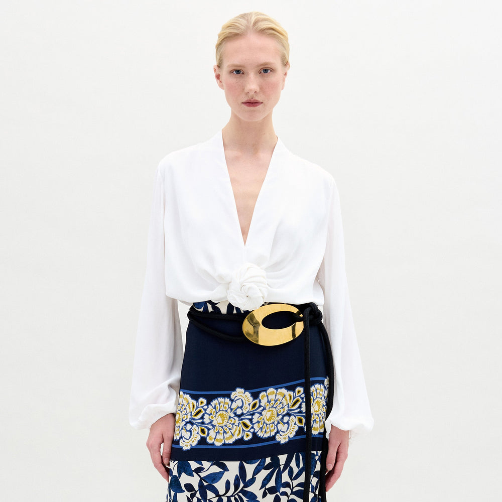 A person with blonde hair wears the SVT DESIGNS Rocio Blouse—white, long-sleeved, with a deep V-neck and flower detail at the waist—paired with a navy skirt featuring yellow and white florals and an asymmetrical hem. The backdrop is plain and light.