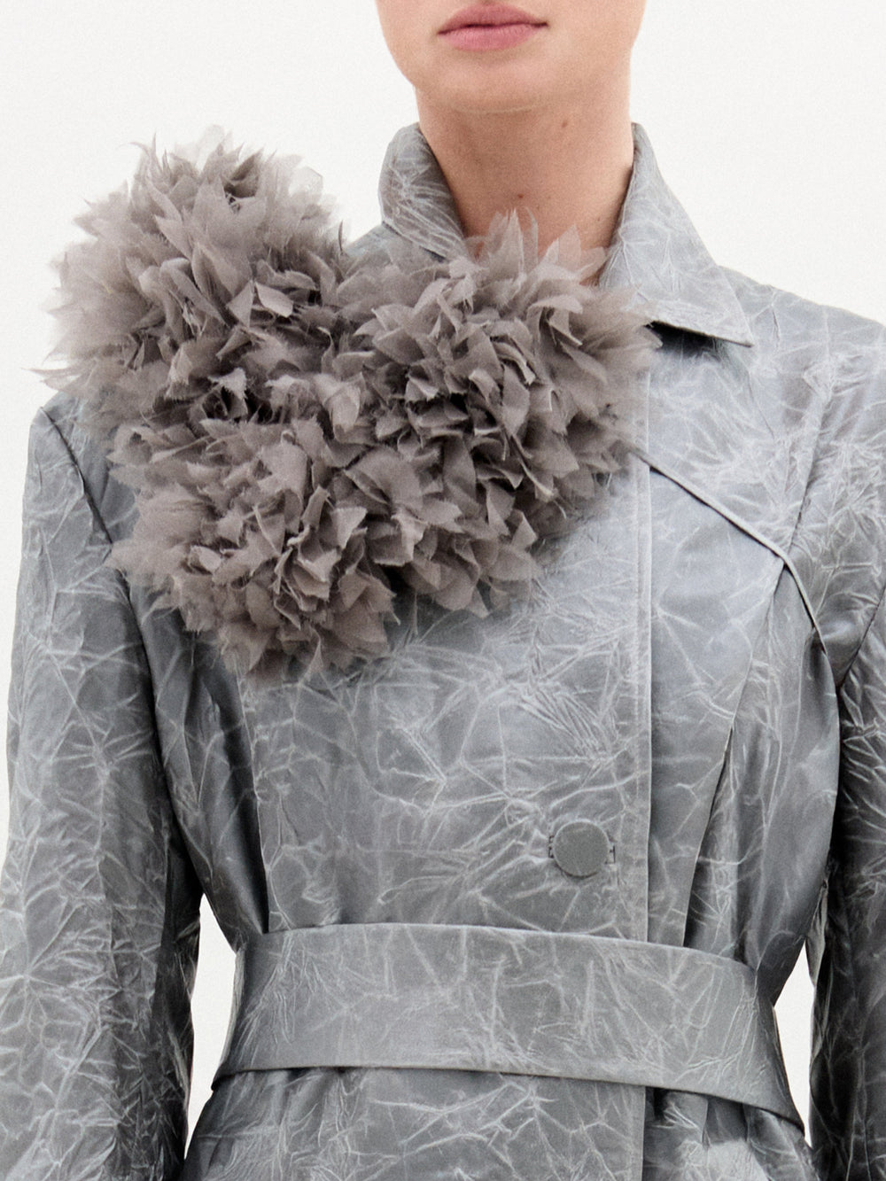A person wears SVT DESIGNS' Romy Collar Piece, a large, fluffy gray floral accessory, pinned on the left shoulder of a light gray Italian-fabric trench coat; only the lower face and upper body are shown.