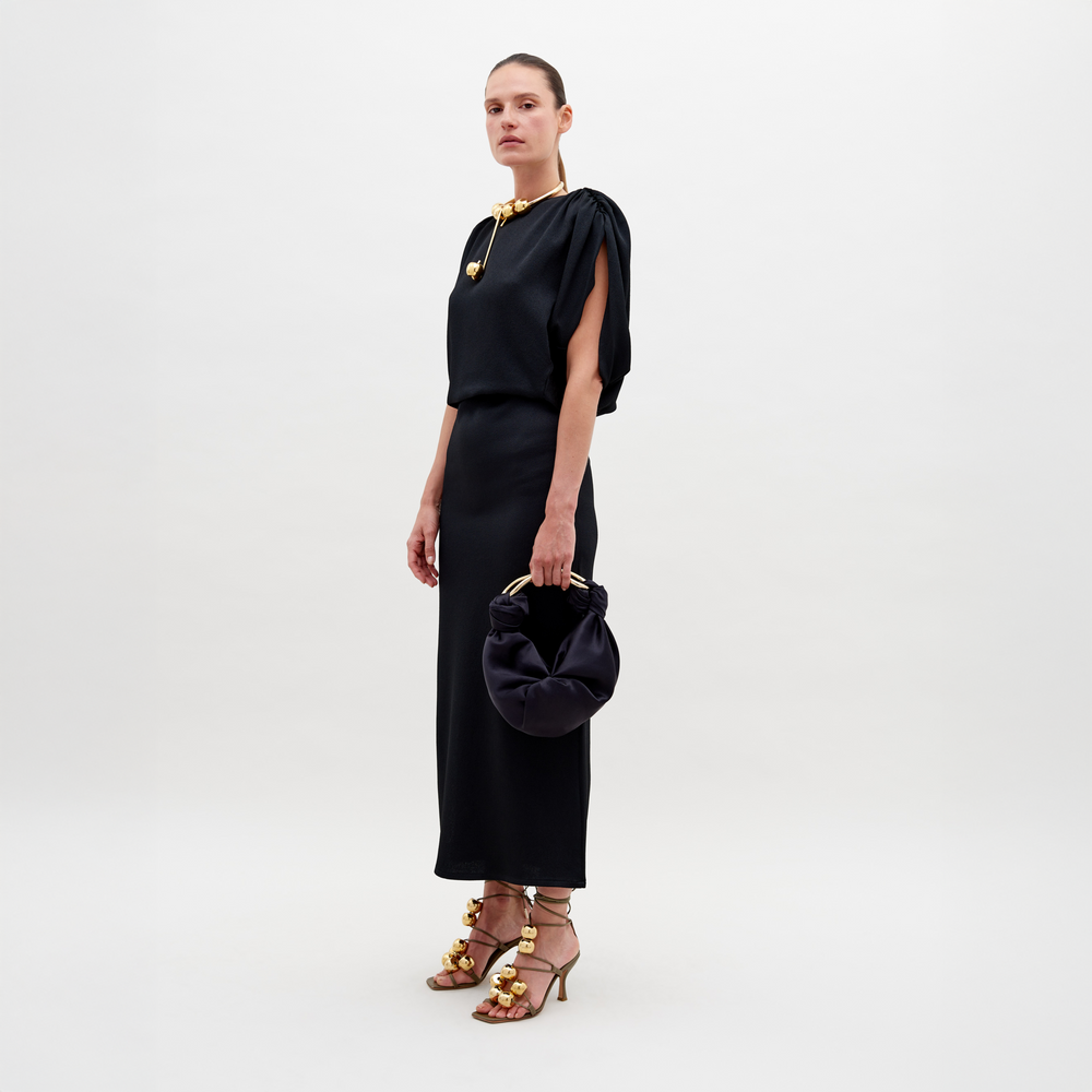 A person showcases the long black Reese Dress by SVT DESIGNS, featuring one sleeve. They hold a dark round handbag, with hair styled back. Gold strappy high-heeled sandals and a gold necklace complete the look against a plain white background.
