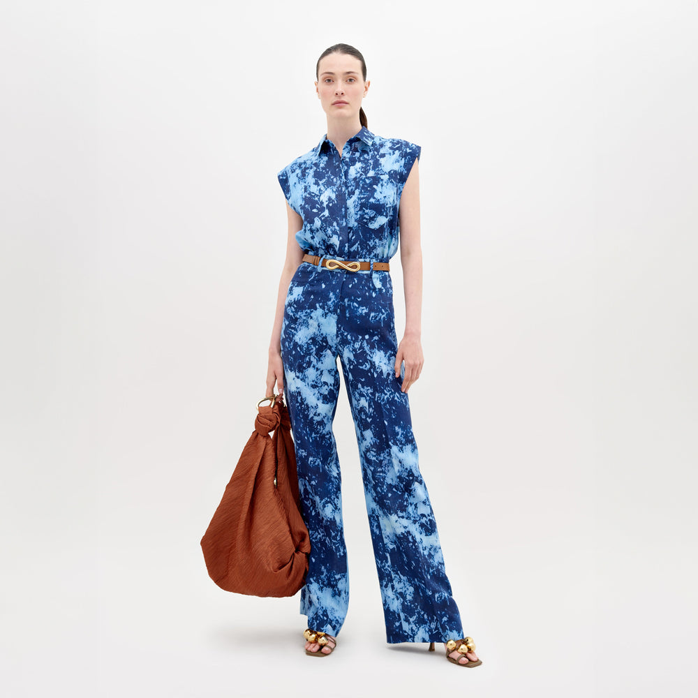A person models the SVT DESIGNS Emine Pant, a blue sleeveless tie-dye jumpsuit with wide-leg trousers and a gold belt. They carry a large brown bag and wear open-toed sandals featuring gold accents, set against a simple light background.