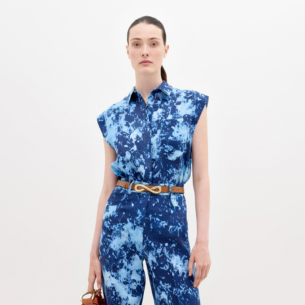 A woman stands against a white background, wearing the SVT DESIGNS Rina Blouse. It features Italian textiles in blue tie-dye, is sleeveless with two chest pockets and a tan belt. She holds a brown handbag and her hair is tied back.