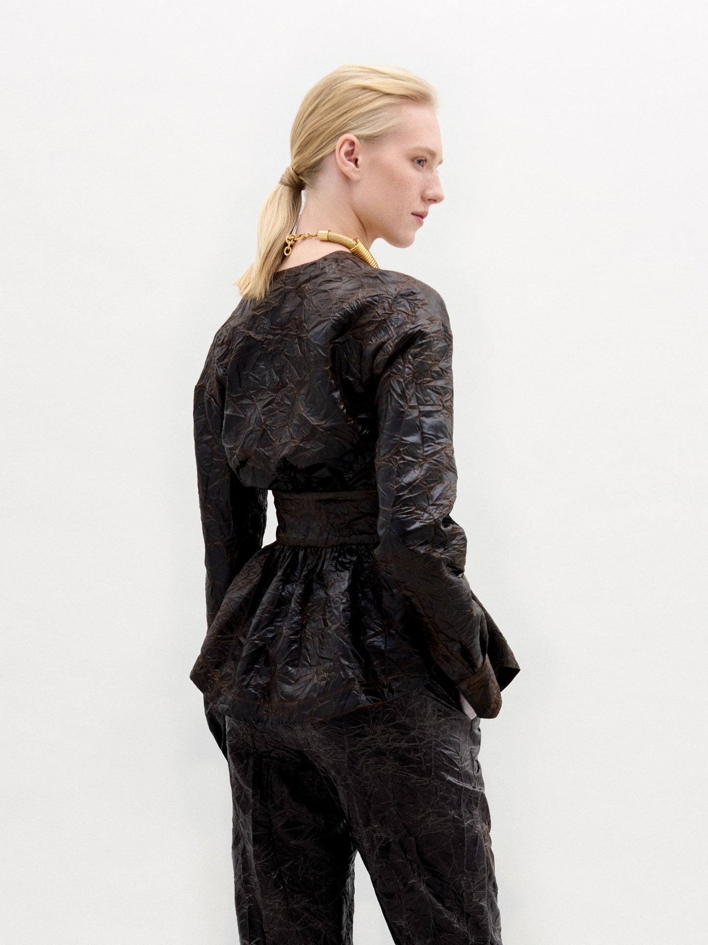 A blonde with a ponytail models the textured dark Saanvi Blouse and matching pants by SVT DESIGNS, featuring Italian fabric and a peplum waist. They're wearing a chunky necklace on a plain white backdrop, turned slightly away from the camera.