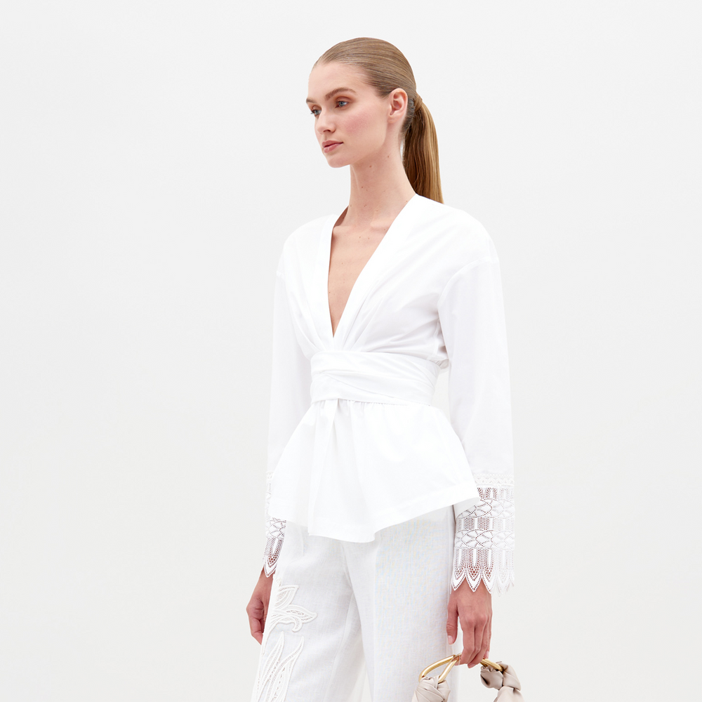 A person with straight, pulled-back hair wears the Saanvi Blouse by SVT DESIGNS—a white cotton top with a deep V-neck and lace-detailed long sleeves—paired with white embroidered pants, holding a small handbag against a plain white background.