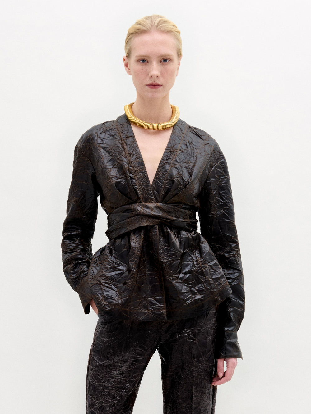 A person with light skin and blonde hair pulled back wears the Saanvi Blouse by SVT DESIGNS—a shiny, dark brown textured blouse with a fitted waist—paired with matching pants and a chunky gold necklace, against a plain white background.