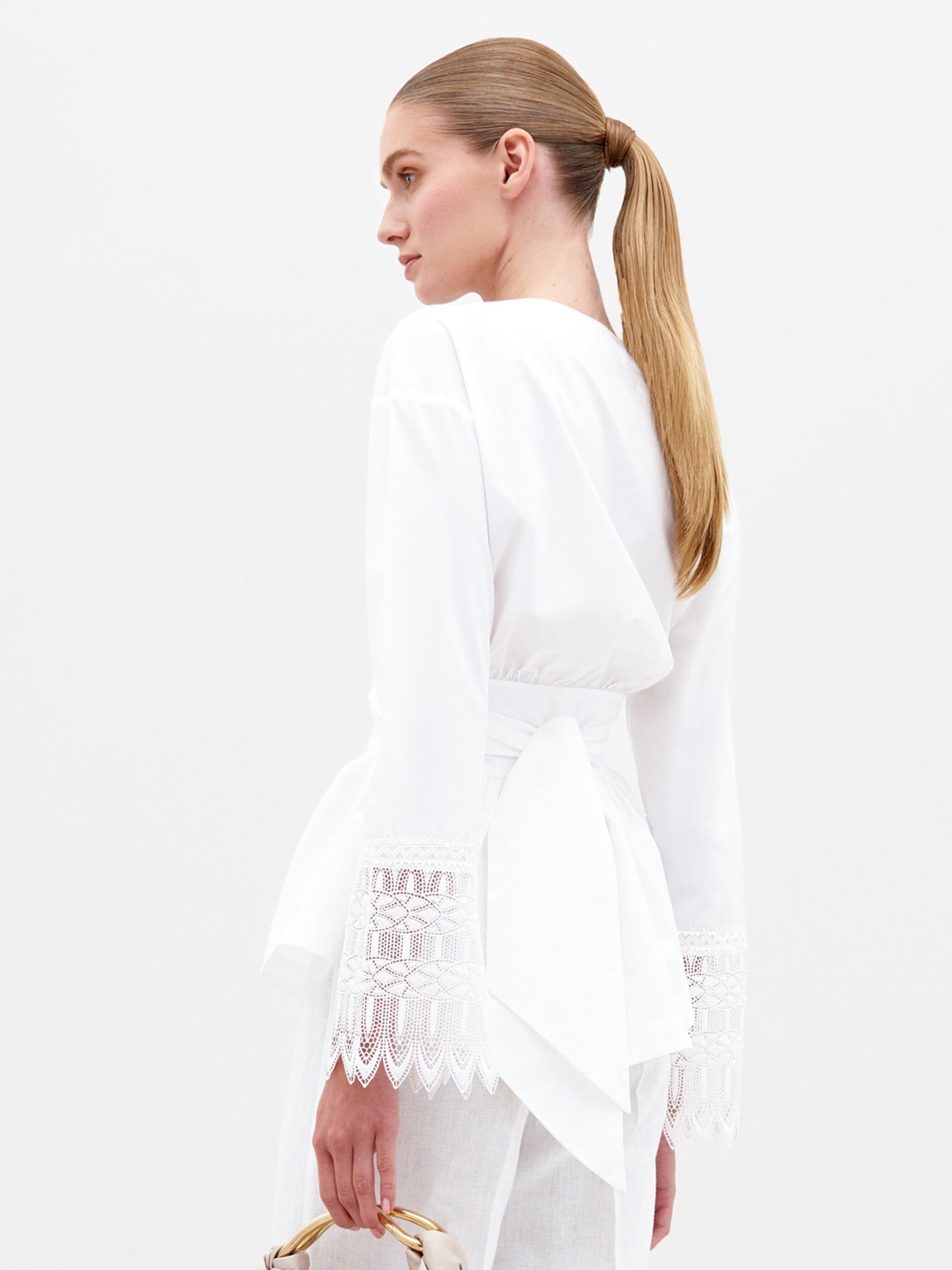 A woman with a sleek ponytail models the Saanvi Blouse—a white cotton top with lace-detailed cuffs and a wide wrap waist tie—while holding a small bag against a plain white background.