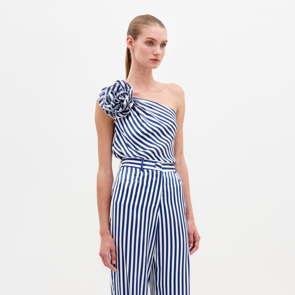 A woman stands against a white background wearing the Sebastiana Blouse by SVT DESIGNS—a one-shoulder, blue and white vertically striped top with an asymmetrical design and large matching floral appliqué on the shoulder. Her hair is pulled back tightly.