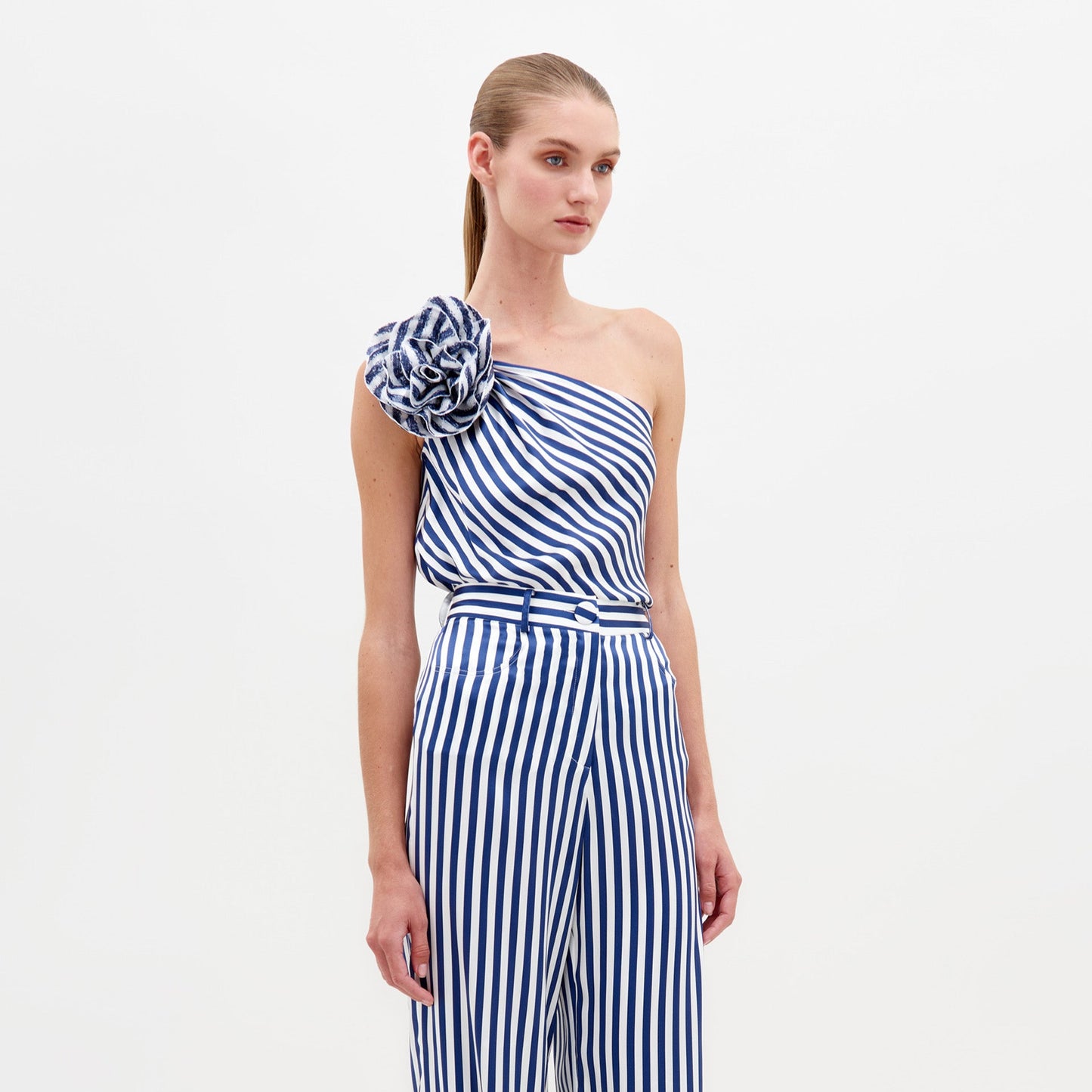 A woman stands against a white background wearing the Sebastiana Blouse by SVT DESIGNS—a one-shoulder, blue and white vertically striped top with an asymmetrical design and large matching floral appliqué on the shoulder. Her hair is pulled back tightly.