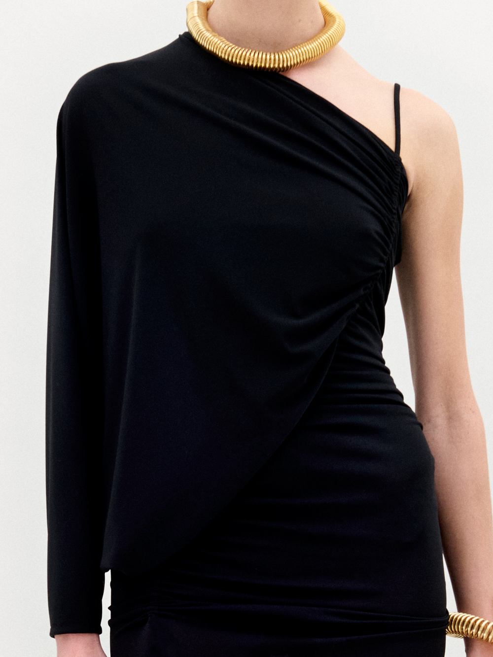 A person wears the Shiri Dress by SVT DESIGNS—a black, asymmetrical one-shoulder column maxi in Italian fabric—paired with a bold gold coiled necklace and bracelet, photographed against a plain light background.