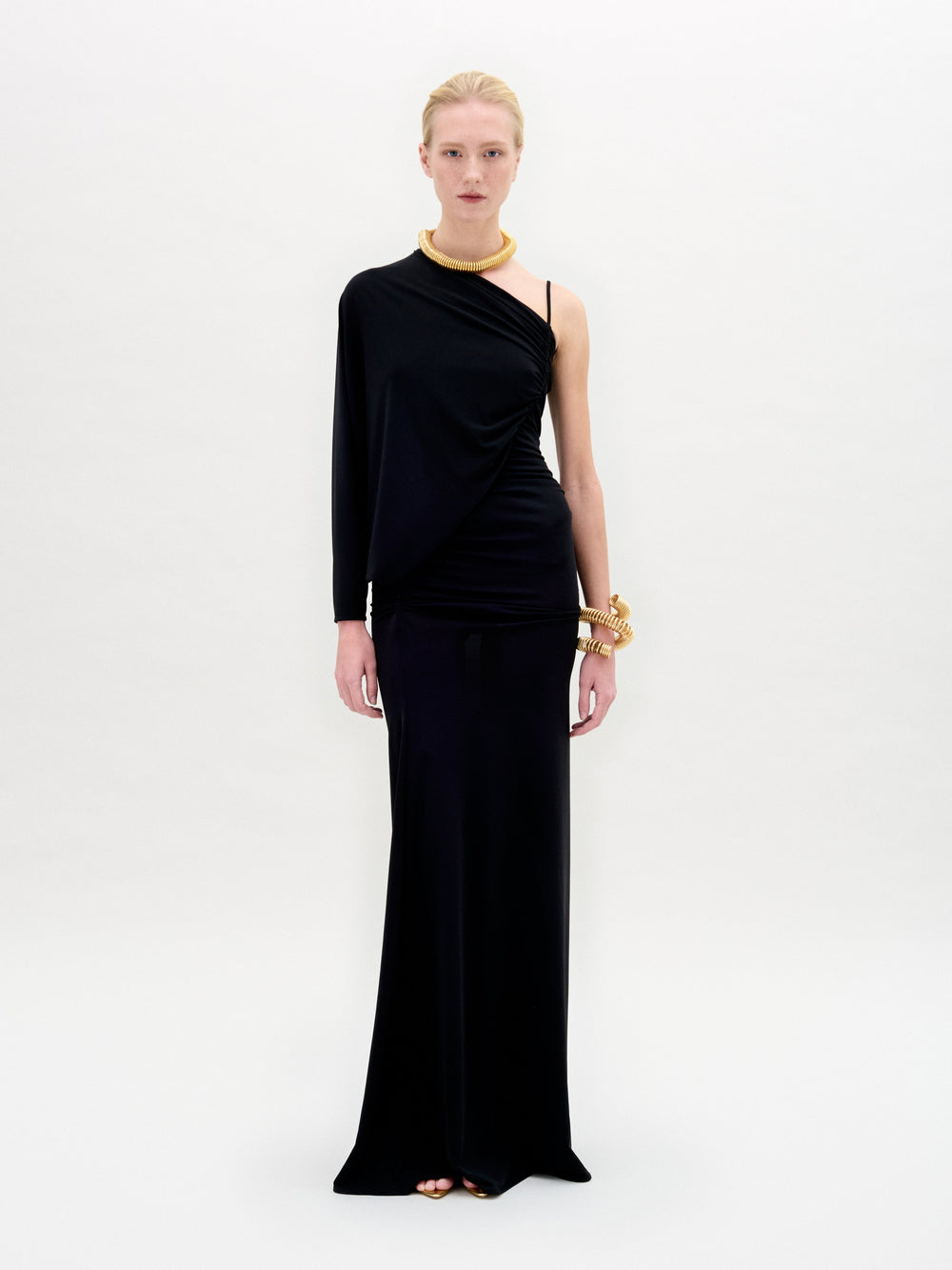 A woman stands against a white background wearing the SVT DESIGNS Shiri Dress, a black one-shoulder maxi with a column silhouette, styled with gold jewelry. She faces forward with a neutral expression.