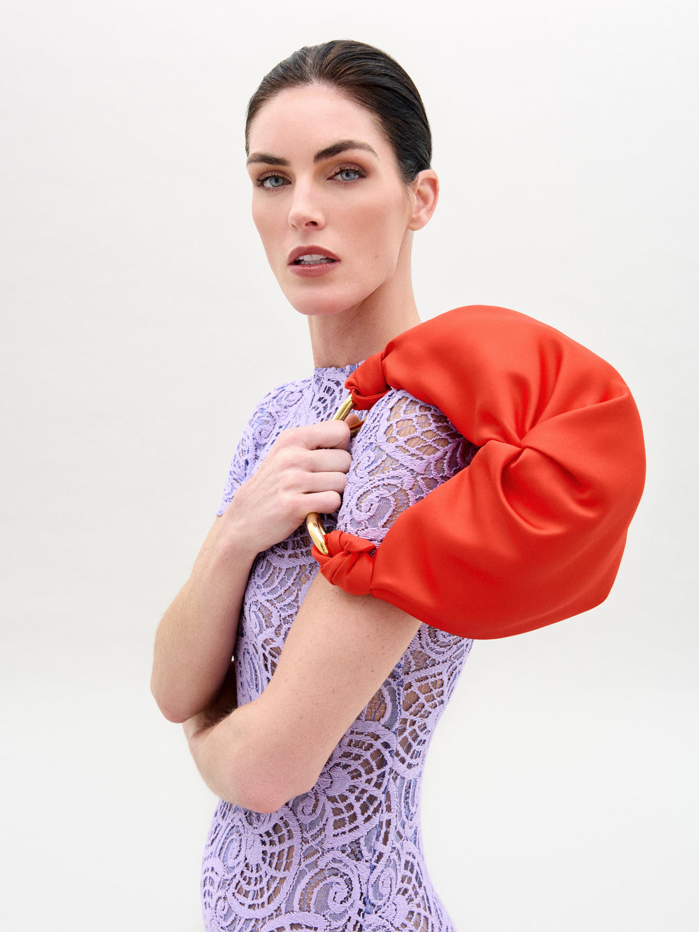 A woman with fair skin and dark hair wears a lavender lace dress and carries the Sofia Bag Small by SVT DESIGNS, a bright orange ruched handbag with a gold-toned handle, over her shoulder against a plain light background.