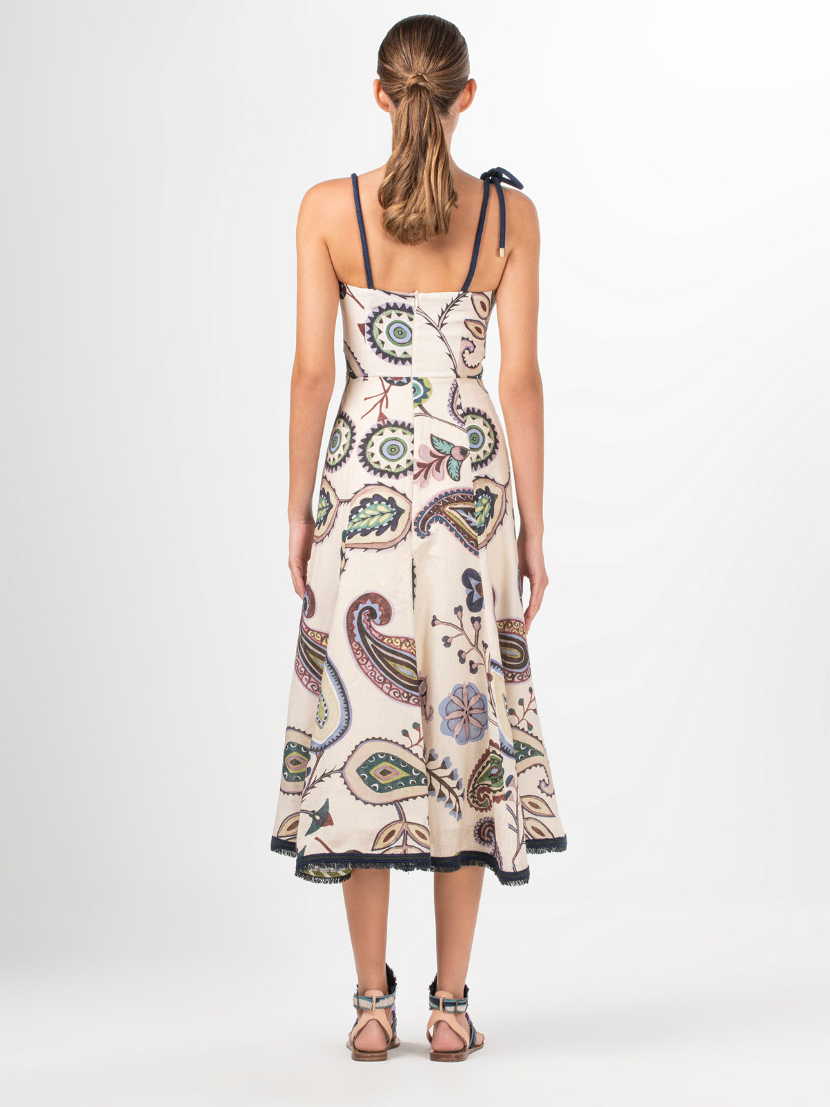 A woman with her hair tied back wears the Dalil Dress by Silvia Tcherassi US. The sleeveless midi dress features paisleys, abstract designs, and fringe lace detailing on the back. She pairs it with flat, open-toed sandals while facing away from the camera.