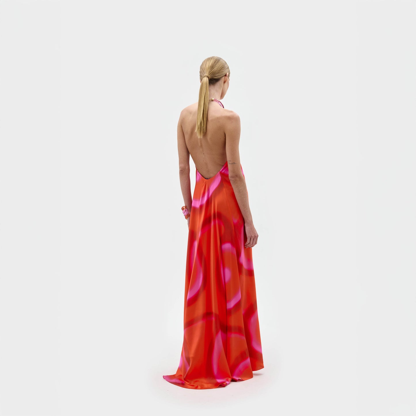 A person with long blonde hair in a low ponytail stands with their back to the camera, wearing the SVT DESIGNS Jesse Dress—a backless, floor-length maxi dress featuring a red and pink abstract print and halter neckline—against a plain white background.