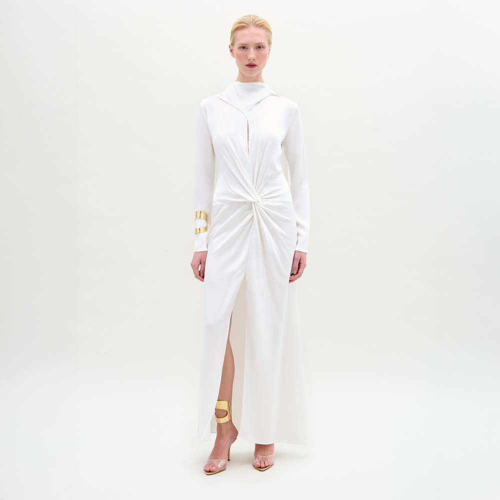 A woman poses against a white backdrop in the SVT DESIGNS Sue Dress, a satin maxi with a central twist and front slit. She pairs it with slicked-back hair, gold cuff bracelets, and open-toe heels featuring gold ankle accents.